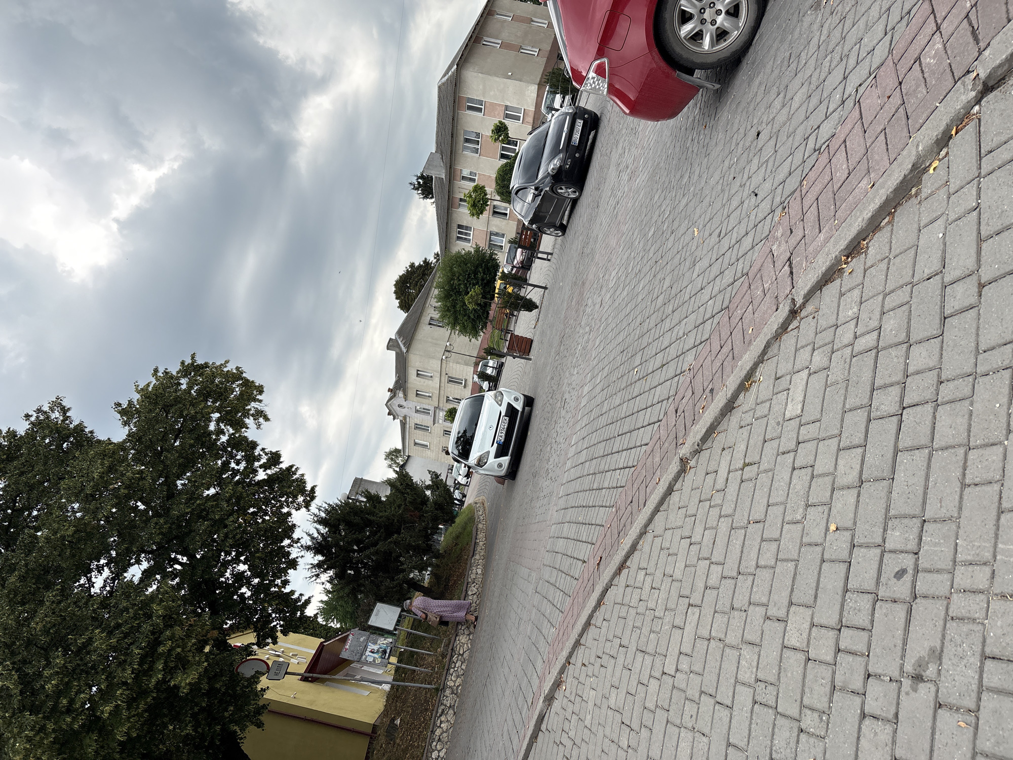 In Lanivtsi, Ukraine, on a cloudy day, a street scene unfolds. Paved with gray interlocking bricks, the street curves gently. A red car occupies the foreground on the right, its rear quarter visible. Further down the street, a black hatchback is parked, and a white sedan is in motion. On the left, a woman in a long, floral dress stands near a yellow building, looking at information boards. The building has a dark-colored awning and a sign with Cyrillic text reading "НЕДІЛЯ" (Sunday). Trees with green foliage are present, and in the background, a multi-story residential building with pale walls and rows of windows stands under a dramatic, overcast sky.