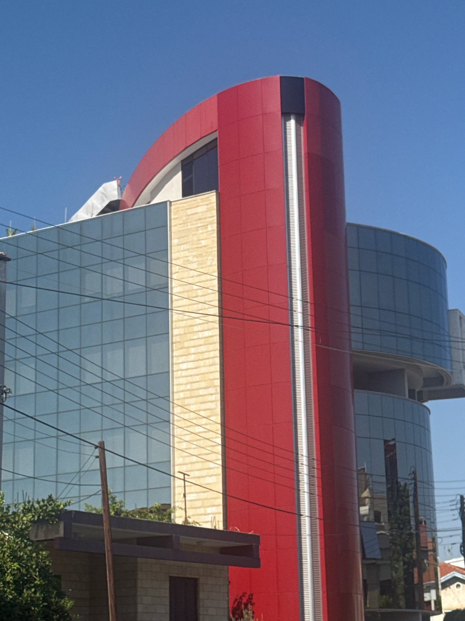 A low-angle, eye-level medium shot captures the striking modern architecture of the Limassol Cooperative Bank in Limassol, Cyprus. The building features a prominent, cylindrical red facade with a vertical metallic stripe. Adjacent to this is a section of the building clad in light-colored stone tiles, and further to the left, a large expanse of blue-tinted glass windows, indicative of office spaces. The building's design incorporates curved elements, as seen in the blue glass sections on the right. Overhead, a clear, bright blue sky serves as the backdrop, with visible overhead electrical wires crisscrossing the scene, suggesting an urban environment. At the base of the building, the roof of a lower structure is visible, partially obscured by lush green foliage. The overall impression is one of contemporary design set against a clear, sunny day in the city.