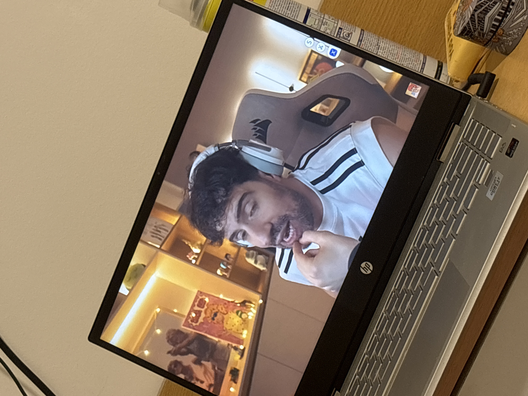 A photograph captures a close-up, angled view of a silver HP laptop displaying a video of a man. The laptop screen is the central focus, showing a man with dark, curly hair and a beard, wearing white over-ear headphones and a white t-shirt with three black stripes on the sleeves, characteristic of Adidas attire. His mouth is slightly open, and he is looking directly towards the camera, with one hand raised near his chin or mouth, suggesting he is actively speaking or reacting.
In the background of the video, a grey gaming chair with a prominent logo (partially visible) is seen, along with a set of shelves behind him. These shelves contain various decorative items and a brightly illuminated strip light along one edge. A framed picture or artwork is also visible on one of the shelves.
At the top right corner of the laptop screen, small circular icons are present, likely part of a video call or streaming interface, including a blue 'X' icon. The physical laptop features a silver keyboard deck and a black bezel around the screen, where an "hp" logo is visible on the bottom left. To the right of the laptop, on a wooden surface, a yellow-capped cylindrical object with text (unreadable) and another patterned object are partially visible. The image is taken indoors, with ambient lighting.