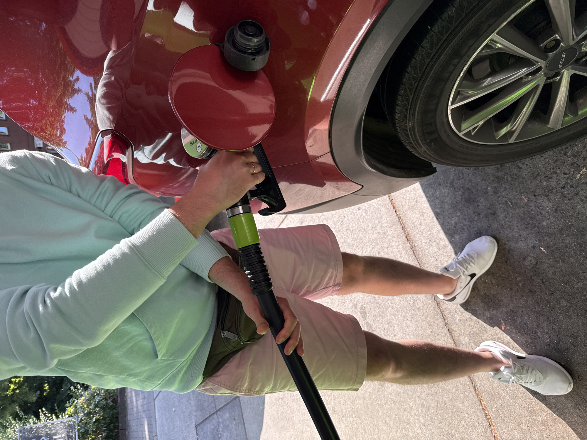 A person is standing next to a red car at a gas station in Bad Lauterberg im Harz, Germany. The person is wearing a light blue hoodie, light pink shorts, and white sneakers. They are holding a black fuel nozzle with a green connector and are in the process of refueling the car. The car is a dark red SUV with alloy wheels. The ground is paved with concrete and there is some greenery in the background, suggesting an outdoor location. The scene is illuminated by sunlight, casting shadows on the ground.