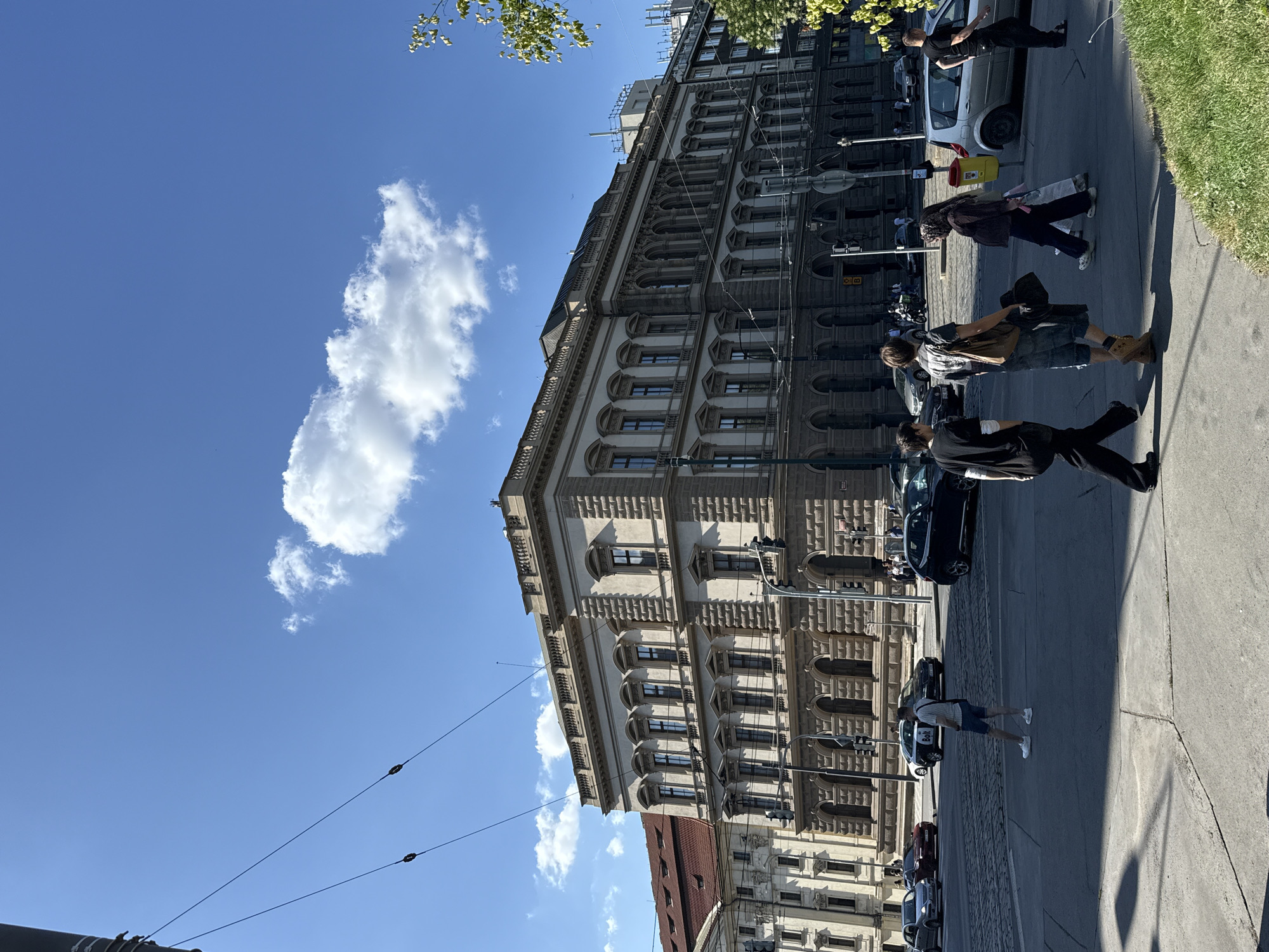 The image depicts a street-level view of a prominent, ornate building in Prague, Czechia, bathed in bright sunlight under a blue sky with scattered white clouds. The building, appearing to be of Neoclassical or Renaissance architectural style, features multiple stories with numerous arched windows and decorative stonework. It dominates the background. In the foreground, a street intersection is visible with paved surfaces and pedestrian crossing markings. Several cars are present on the road, including a silver sedan, a dark-colored car, and a white Bolt branded vehicle. A traffic light is visible on a pole.
In the middle ground and foreground, several pedestrians are crossing the street. One man in a light-colored shirt and dark shorts walks towards the viewer's right. Another group of three individuals are crossing in the opposite direction; one wearing a black top and black pants, another in a patterned shirt and denim shorts carrying a brown bag, and a third person in a dark headscarf and dark pants. To the far right, another man in a black t-shirt and black pants walks away from the viewer. The scene suggests a busy urban environment in Prague.