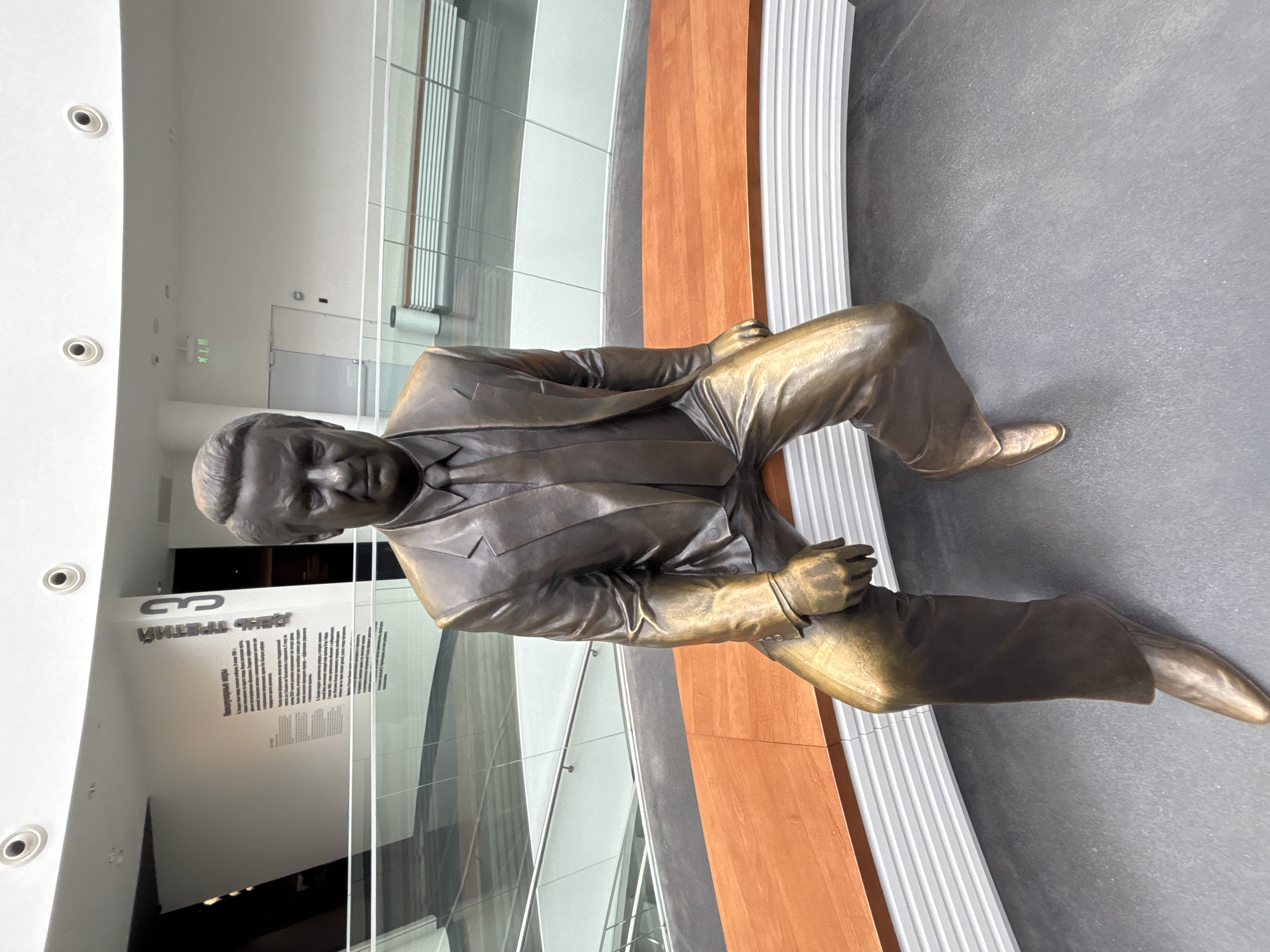 A full-body bronze statue of Boris Yeltsin, the first President of Russia, is depicted seated on a curved wooden bench with white ribbed supports. Yeltsin wears a suit and tie, with his right hand resting on his right thigh and his left hand on the bench beside him; his left leg is slightly extended. The statue is situated on a dark grey floor within the modern, multi-level interior of the Boris Yeltsin Presidential Center (Yeltsin Center) in Yekaterinburg, Russia. The background features white walls, glass and metal railings, and a prominent exhibit on a pillar displaying Cyrillic text. Visible phrases include "ДЕНЬ ТРЕТИЙ" (DAY THREE) and "Непопулярные меры" (Unpopular measures), indicating a museum exhibit. Recessed lighting is visible in the ceiling, and in the distance, light-colored doors, a green exit sign, and a grey waste bin are present.