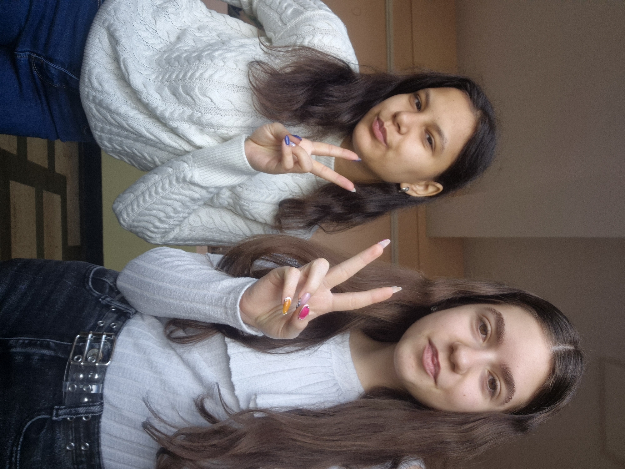 Two young women are posing for a photo. The woman at the top is wearing a white sweater and blue jeans, and she is making a peace sign with her hand. Her fingernails are painted blue. The woman at the bottom is wearing a white top, black pants with a silver belt, and she is also making a peace sign with her hand. Her fingernails are painted in various colors. The background appears to be a wall with a neutral color. The image does not provide information about the city of Pavlikeni, Bulgaria.