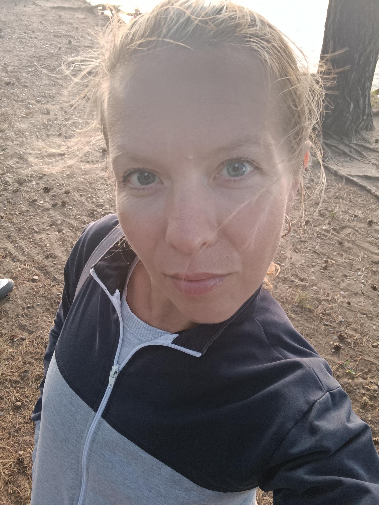 The media file is a selfie of a woman with blonde hair and blue eyes, looking directly at the camera. She is wearing a two-toned zip-up jacket, dark blue on top and grey on the bottom, over a light grey t-shirt. The background suggests an outdoor setting, likely near a body of water, with sandy or gravelly ground covered in pine needles and small cones. A tree trunk is visible to the right of the woman. The lighting indicates it is either early morning or late afternoon, with the sun casting a warm glow. The woman's hair is slightly windswept. The scene appears relaxed and casual. No text is visible. The location context is Chaikovsky, Russia.