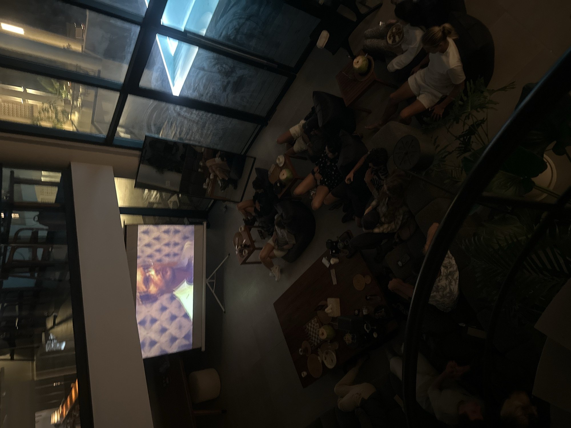 The image depicts an indoor gathering where several individuals are casually watching a projected movie. The room appears to be a spacious, modern living area with a projector screen displaying a man's face. People are seated on beanbag chairs and the floor, some with their legs extended. There are coffee tables with drinks and snacks scattered around. Large windows reveal an outdoor area with a pool, suggesting a contemporary home or a rented villa. The lighting is dim, creating a relaxed, nighttime ambiance. The location is identified as Mengwi, Indonesia, indicating a potentially tropical or subtropical setting.