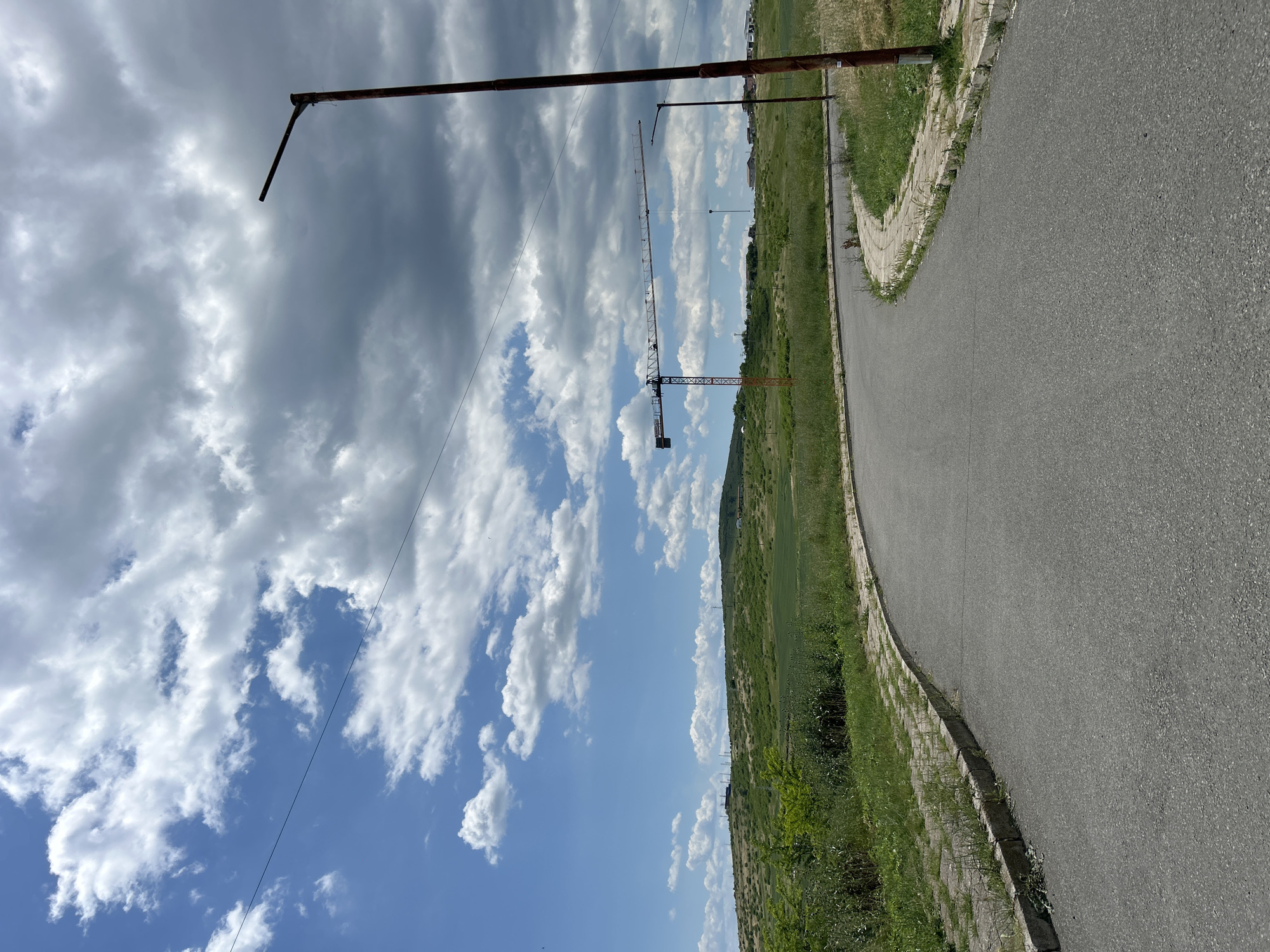 The image depicts a road curving to the right, bordered by grass and a low concrete barrier. In the background, a field stretches towards a distant treeline. Above the field, a construction crane is visible against a partly cloudy sky. The presence of the crane and the field suggest an area of ongoing development. The image is likely taken in an urban-fringe area. Given the prompt mentions the city of Burgas, Bulgaria, the location is likely on the outskirts of Burgas where new construction meets agricultural land. Overhead wires are visible, possibly power lines. A rusting metal pole with a short, angled arm is in the foreground.