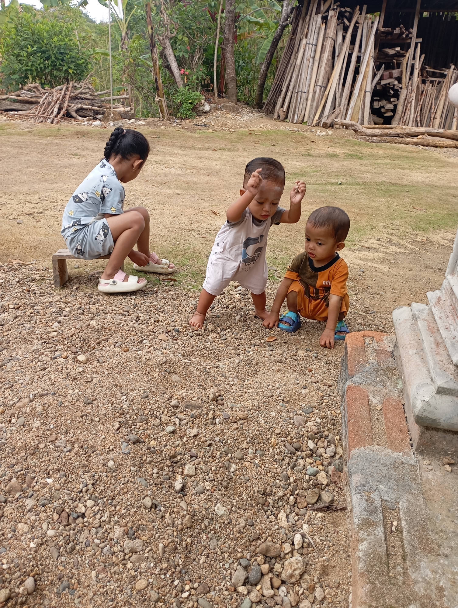 In Ketepung, Indonesia, three young children are playing on a dirt and gravel ground. A young girl with braided hair, wearing a light blue pajama set with a cartoon pattern, is sitting on a small wooden bench. To her right, two younger boys are on the ground. One boy, in a white onesie, appears to be dancing or playing energetically, with his arms raised. The other boy, wearing an orange and brown outfit, is crouched down, observing his surroundings. The environment suggests a rural or semi-rural setting, with a backdrop of trees, vegetation, and a pile of wooden logs. A stone structure with brick accents is visible in the foreground.