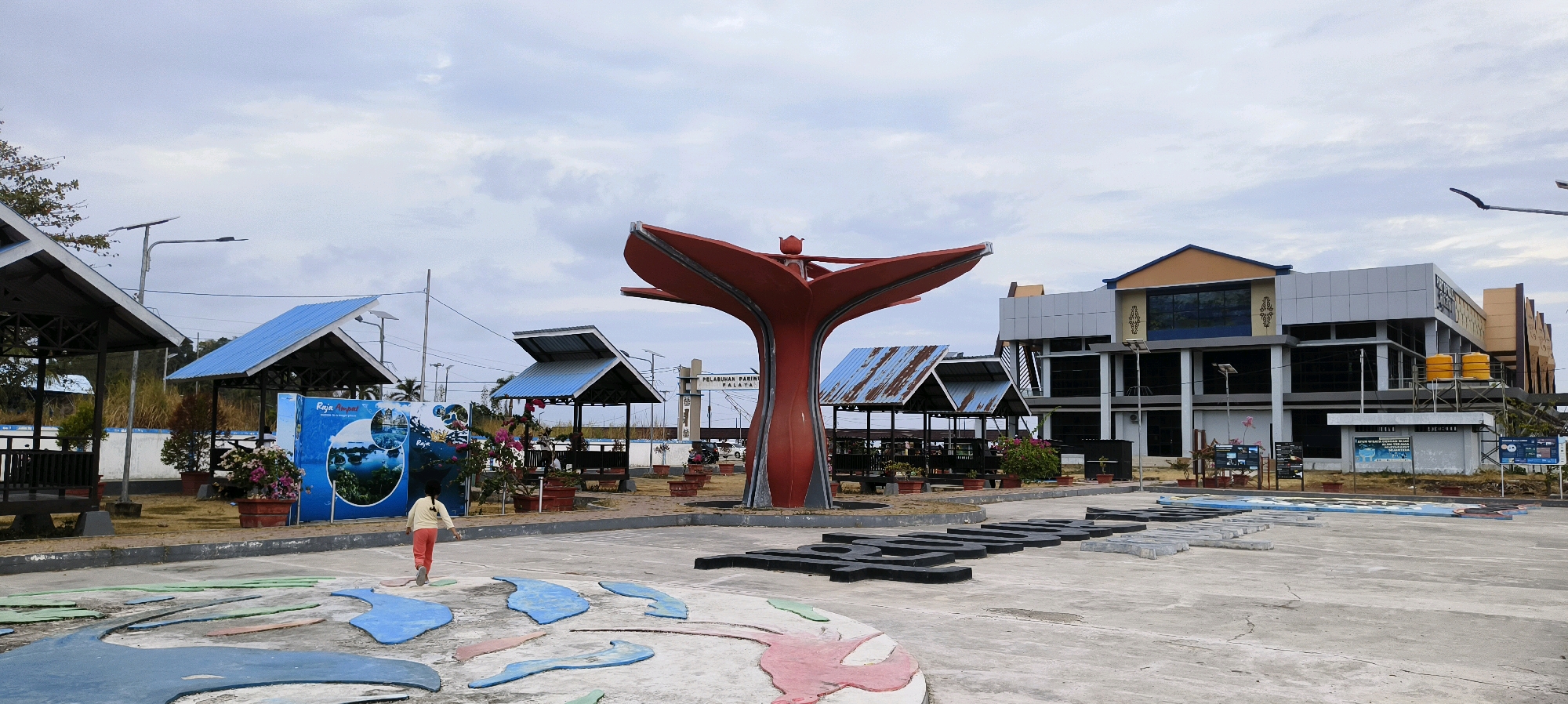 The image depicts the waterfront area of Waisai, Indonesia, likely a public space near the port. Dominating the foreground is a large, red, abstract sculpture resembling an open flower or a stylized bird. In front of this sculpture, a child wearing a light-colored top and orange pants walks away from the camera on a concrete surface decorated with colorful, abstract shapes that resemble a map or geographical features.
Behind the sculpture and the child, several covered structures with blue roofs and open sides are visible, likely shelters or seating areas. A large, blue and white informational poster is propped up near one of these structures, displaying images of marine life and landscapes, with text that includes "Raja Ampat." Further in the background, a modern, multi-story building with a grey and tan facade stands. This building has several windows and appears to be a public facility, possibly related to the port or tourism. Yellow water tanks are visible on a platform attached to the building. The sky is overcast and cloudy, suggesting a diffused light. The environment appears to be a paved public area with some vegetation and trees in the distance. The overall scene conveys a sense of a developing tourist or transportation hub.