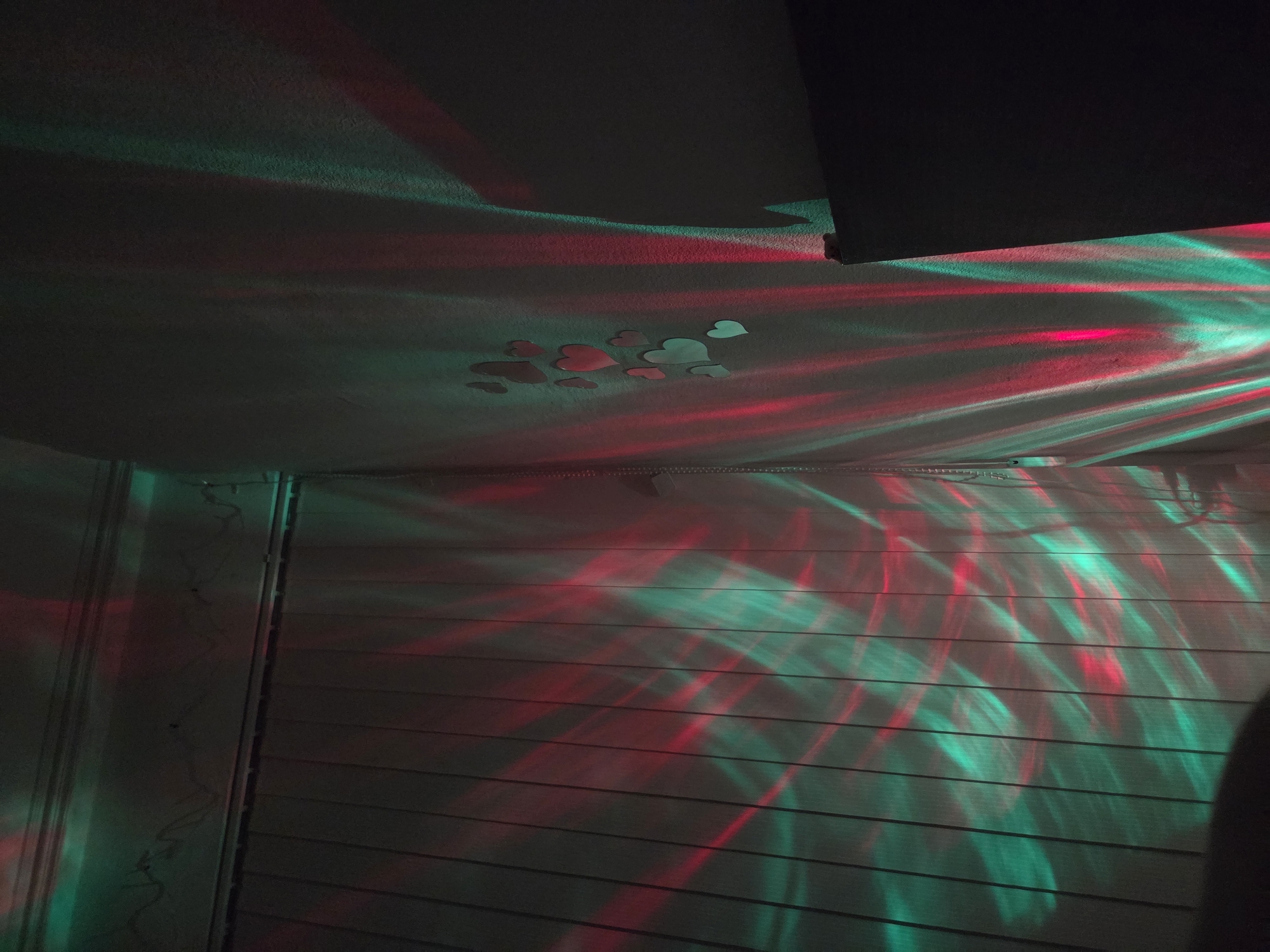 The image shows a corner of a room with a wall and a ceiling. The wall is made of horizontal planks. The ceiling has a rough texture and several heart-shaped decorations attached to it. Red and green light beams illuminate the wall and ceiling, creating a pattern of alternating colors. There is no information about the location being in Pleven, Bulgaria. No people, subjects, actions, or interactions are visible.
