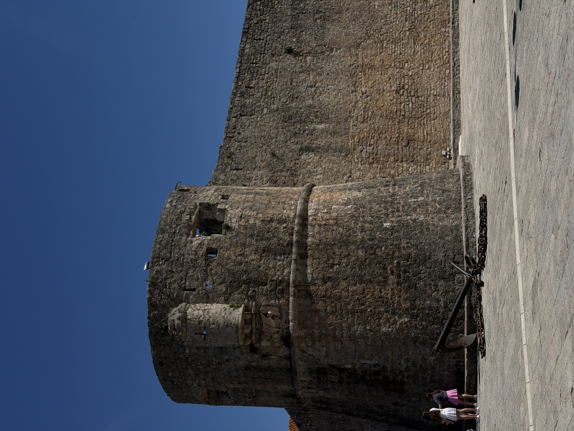 A bright, sunny daytime scene captures a section of a historic stone fortification, likely part of the ancient city walls of Budva, Montenegro. The image, correctly oriented, shows a towering stone wall on the left, characterized by rough, irregular blocks and varying shades of brown and grey, suggesting age. A prominent cylindrical tower or bastion, built into the main wall, extends towards the viewer's lower left, featuring small rectangular openings or windows. One section of the tower reveals a carved stone relief or sculpture.
To the right of the stone wall is a light-colored, paved walkway or plaza, also made of stone or concrete, indicating a well-maintained public area. Strong sunlight casts distinct shadows, suggesting the sun is high in the sky. A large, dark, metal anchor with chains is displayed on the paved ground, positioned close to the wall, hinting at the city's maritime history.
A few individuals are visible on the paved area, appearing to walk or stand near the base of the wall. One person wears a pink skirt and a dark top, while another is in a white top. Their presence gives a sense of scale to the massive fortification. The sky above is a clear, deep blue, with no clouds, reinforcing the impression of good weather. No discernible text is visible in the image. The scene evokes a sense of historical grandeur in a Mediterranean setting.