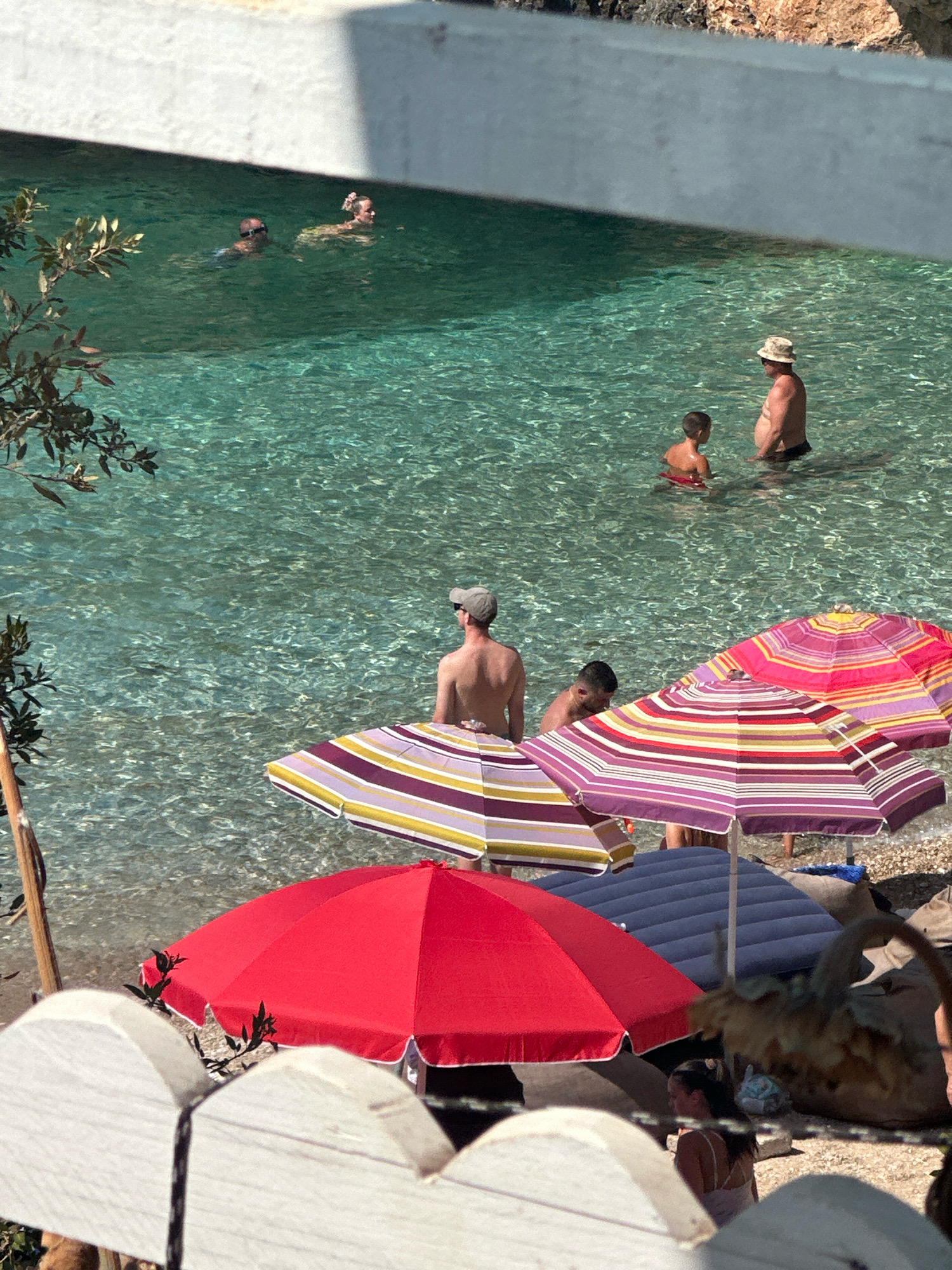 The image captures a sunny day at a beach with clear, turquoise water. Several people are visible in the water, some swimming and others standing. On the shore, a variety of colorful beach umbrellas provide shade. A woman is seated in the foreground, partially obscured by a white fence. In the water, a man in a hat and swimming trunks stands with a child. Further out, two people are swimming. The scene is set against a backdrop of rocky cliffs. The overall impression is one of a leisurely day at a picturesque beach.