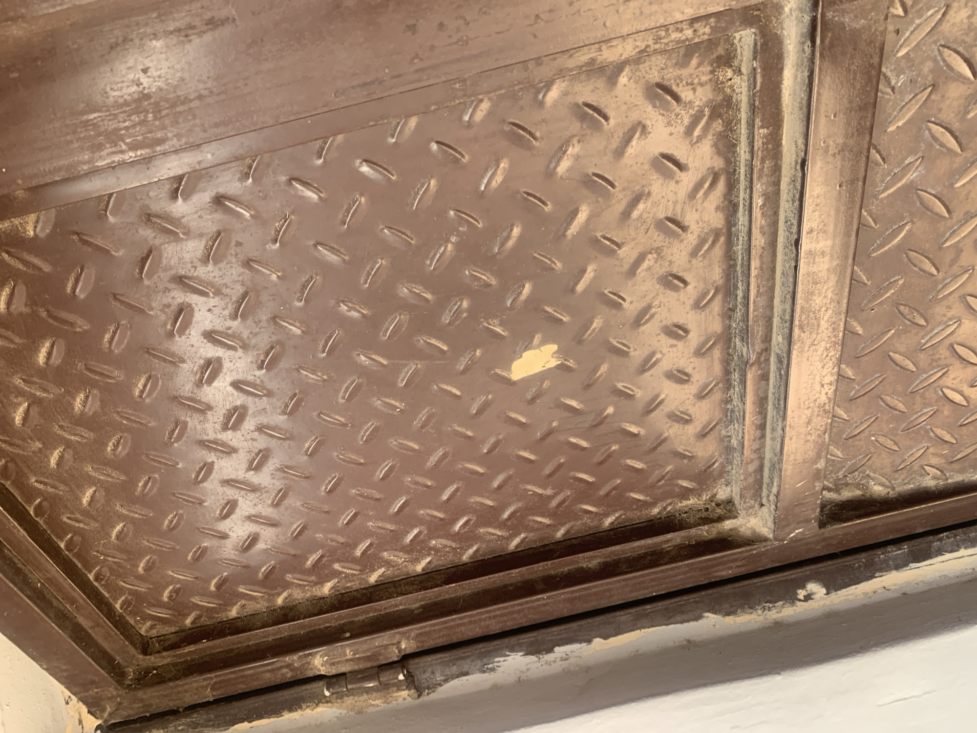 The image shows a close-up view of a textured metal surface, likely a door or gate, with a diamond plate pattern. The metal is painted a dark brown, and it appears weathered, with some scuff marks and dust accumulation. A small, discolored patch, possibly a sticker or paint chip, is visible on the upper right quadrant of the patterned area. The metal framing surrounding the patterned section is also visible, as is a portion of a light-colored wall to the left. The lighting suggests an indoor or shaded outdoor environment. The location is Jauro Yinu, Nigeria.