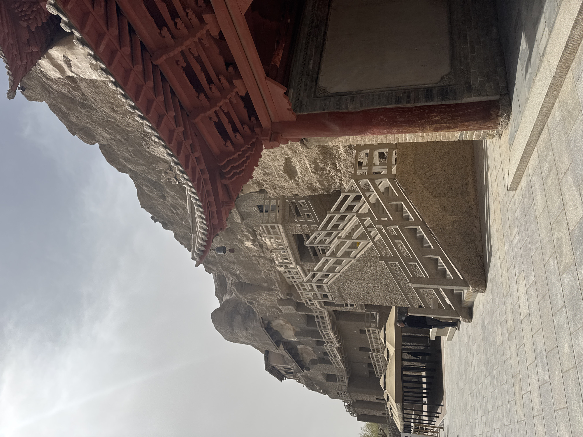 This photograph shows the Mogao Caves, located in Dunhuang City, China. A red, ornately designed wooden structure is at the top of the image, partially obscuring the view of a tall, rocky cliff face that forms part of the cave complex. The cliff face features a series of square openings and a white, angular framework, possibly scaffolding or a pathway, constructed along the cliff. To the right of the cliff face is a stone wall or platform. The sky is light gray. The photograph appears to be taken from below.