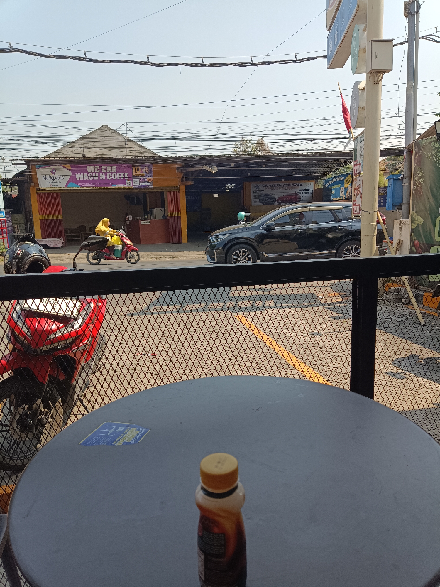 The image captures a street scene in Jember Kidul, Indonesia, viewed from an elevated perspective, possibly from a cafe or restaurant. In the foreground, a dark circular table with a bottle of iced coffee is visible, along with a portion of a red motorcycle parked behind a metal mesh fence.
The midground shows a street with several vehicles, including a black SUV and a red scooter ridden by a person wearing a yellow hijab and outfit. The scooter has a yellow container strapped to its back. Adjacent to the street is a building with a prominent sign that reads "VIC CAR WASH N COFFE" and "MyRepublic #WinnerBaik". The building appears to be a car wash and coffee shop.
The background reveals a clear sky, overhead power lines, and more buildings. The overall environment suggests a sunny day in a commercial area of the city.