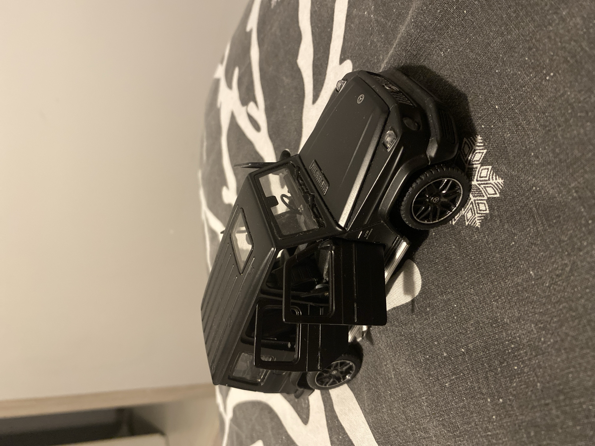 The image shows a black toy Mercedes-Benz G-Class SUV with its doors open, placed on a patterned surface. The toy car is detailed, featuring visible rims, headlights, and a sunroof. The background is a plain, light-colored wall. The surface the car is on appears to be a fabric with a black and white abstract pattern. No specific location in Łódź, Poland, is identifiable from the image.