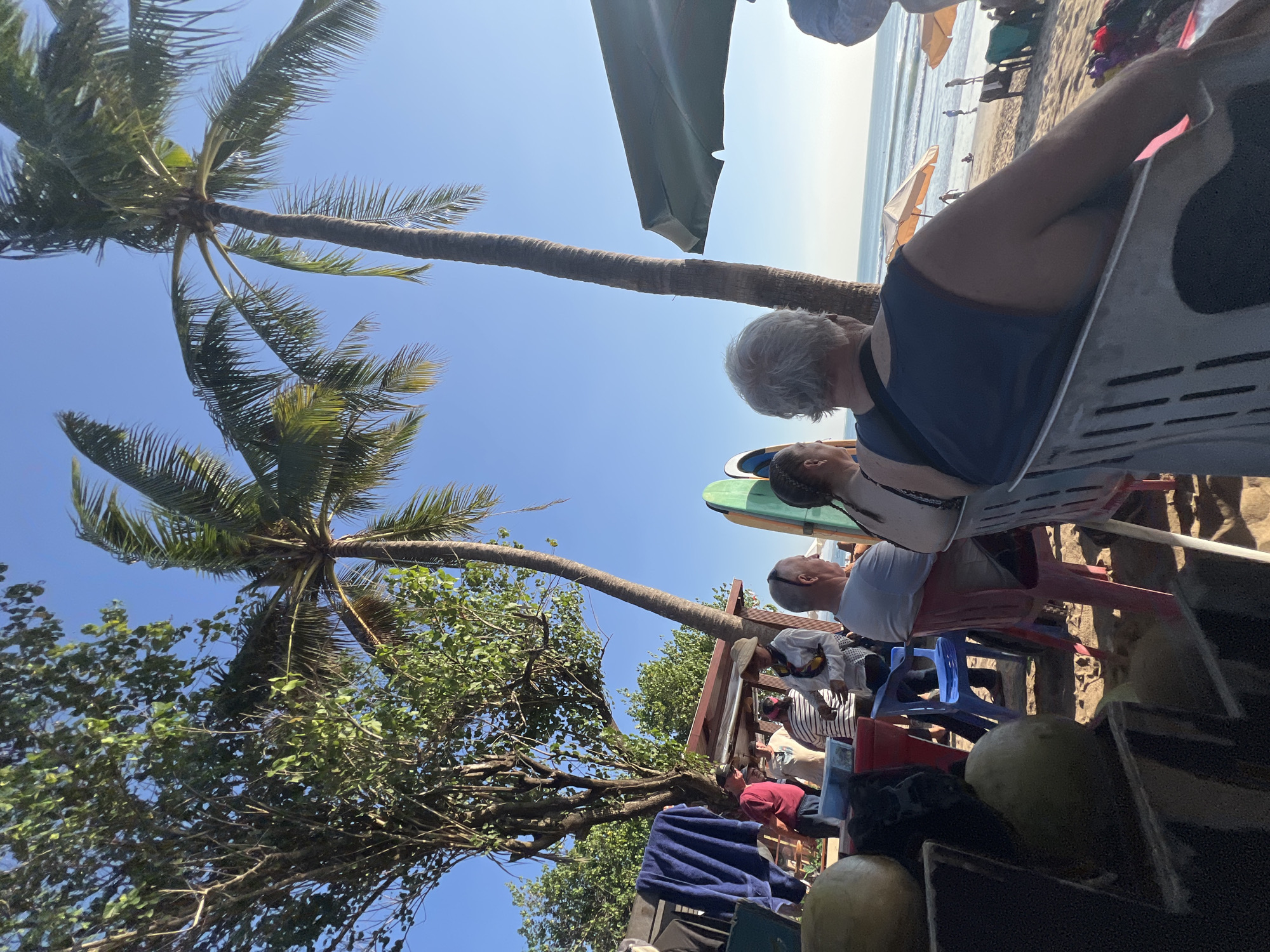 In Kuta, Indonesia, this scene captures a relaxed beachside gathering. Several people are seated at tables under palm trees, some facing the ocean, others conversing. Surfboards are visible near a food stall. In the foreground, a few large green fruits are arranged on a shelf. The ocean and a clear blue sky form the backdrop. The atmosphere appears casual and leisurely, typical of a beach resort.