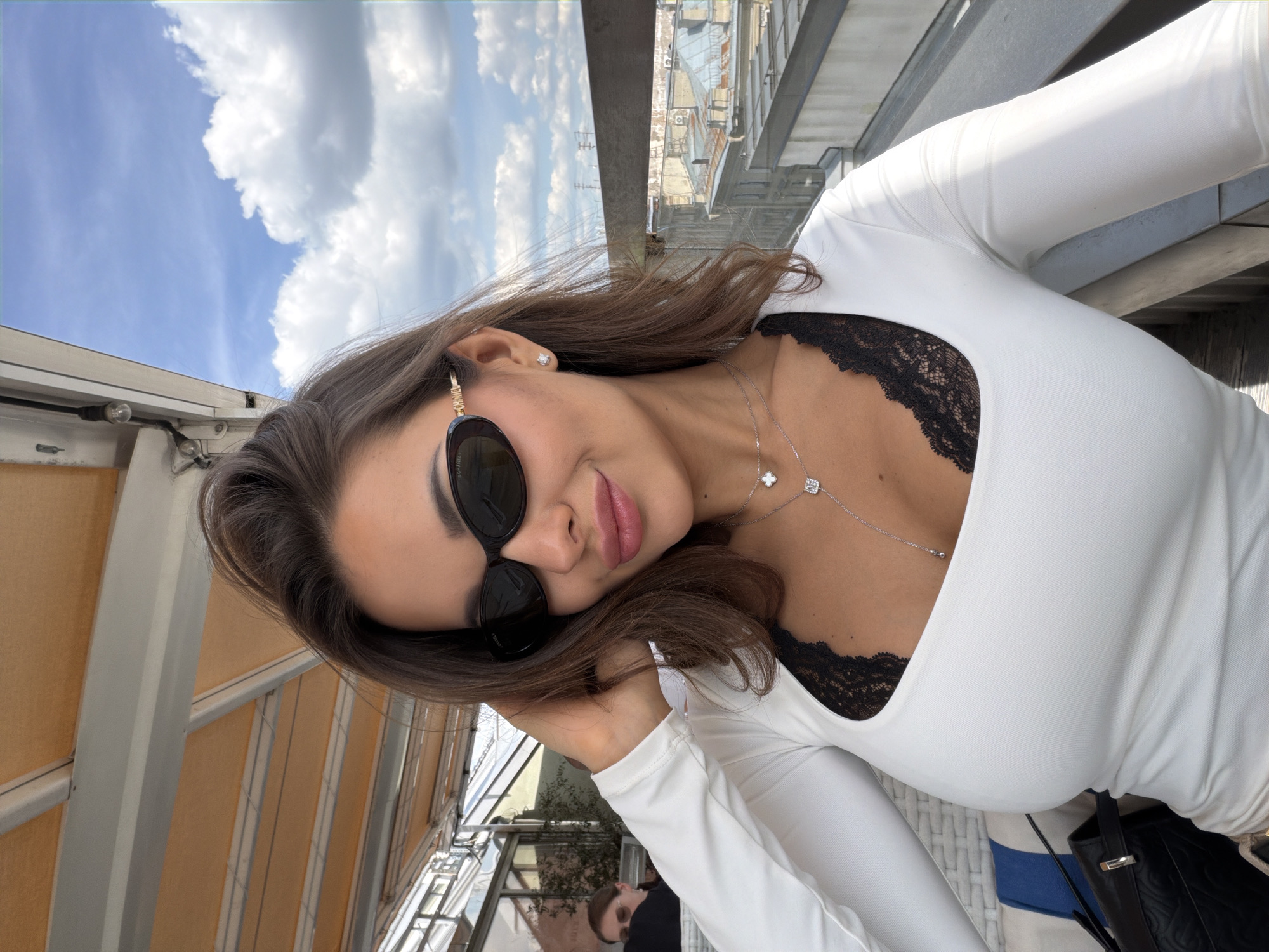 A young woman with dark hair, wearing sunglasses and a white long-sleeved top with a black lace trim, is taking a selfie on a rooftop terrace in Saint Petersburg, Russia. She is leaning her head to the side and touching her hair with her right hand, and she is wearing a necklace with two pendants. Behind her, the sky is blue with white clouds, and a cityscape with several buildings can be seen. The terrace has a retractable awning overhead and a railing in the foreground. Another person, partially visible, is seated at a table in the background.