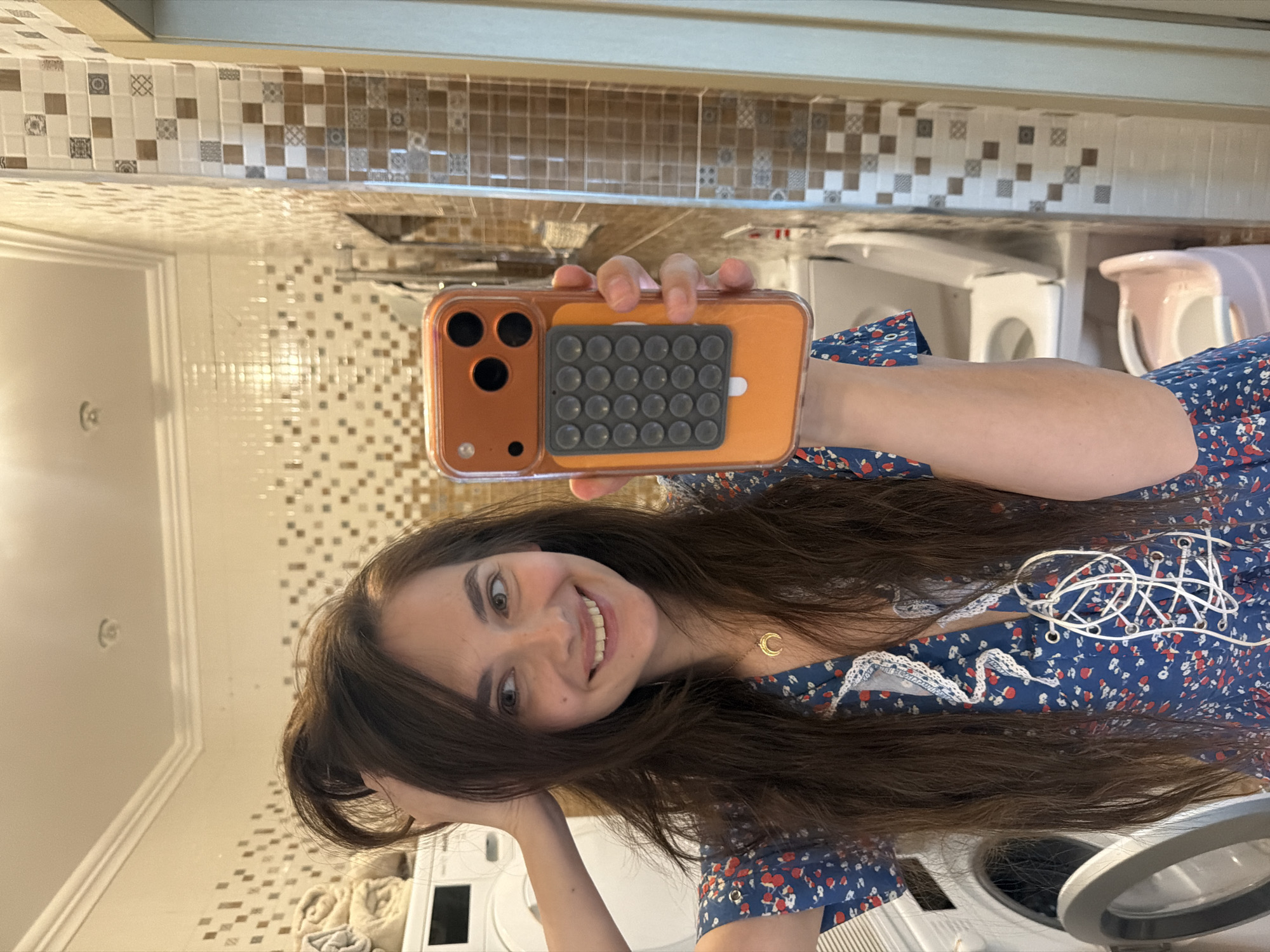 The image depicts a young woman with long, dark brown hair, taking a mirror selfie in a bathroom. She is wearing a blue dress with a floral print and a white lace trim. Her left arm is raised, holding her hair up, while her right hand holds an orange smartphone with a pop socket attached to the back. The bathroom appears to have tiled walls with a mosaic pattern of brown and cream squares. In the background, a white washing machine and what appears to be a toilet with a child's potty seat are visible. The overall scene is casual and domestic. There is no information in the image to suggest this location is in the city of Ufa, Russia.