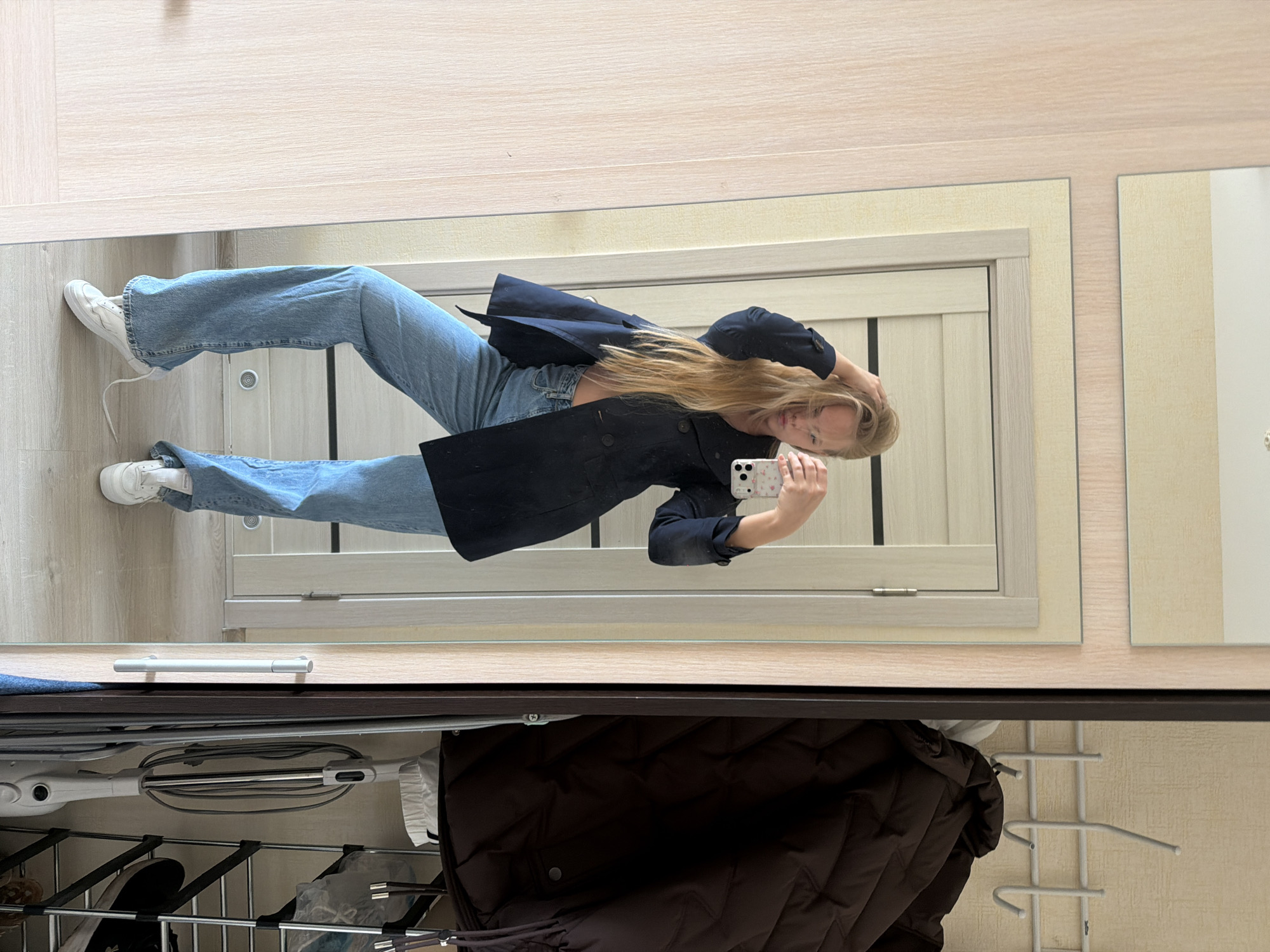 A woman is standing in front of a mirror, taking a selfie with her smartphone. She is wearing light blue jeans, white sneakers, and a navy blue blazer. Her blonde hair is long and loose. The mirror is mounted on a wall above a storage unit. Below the mirror, there is a rack with clothing and other items, including a vacuum cleaner. The room appears to be indoors, possibly in a residential apartment in Izhevsk, Russia.