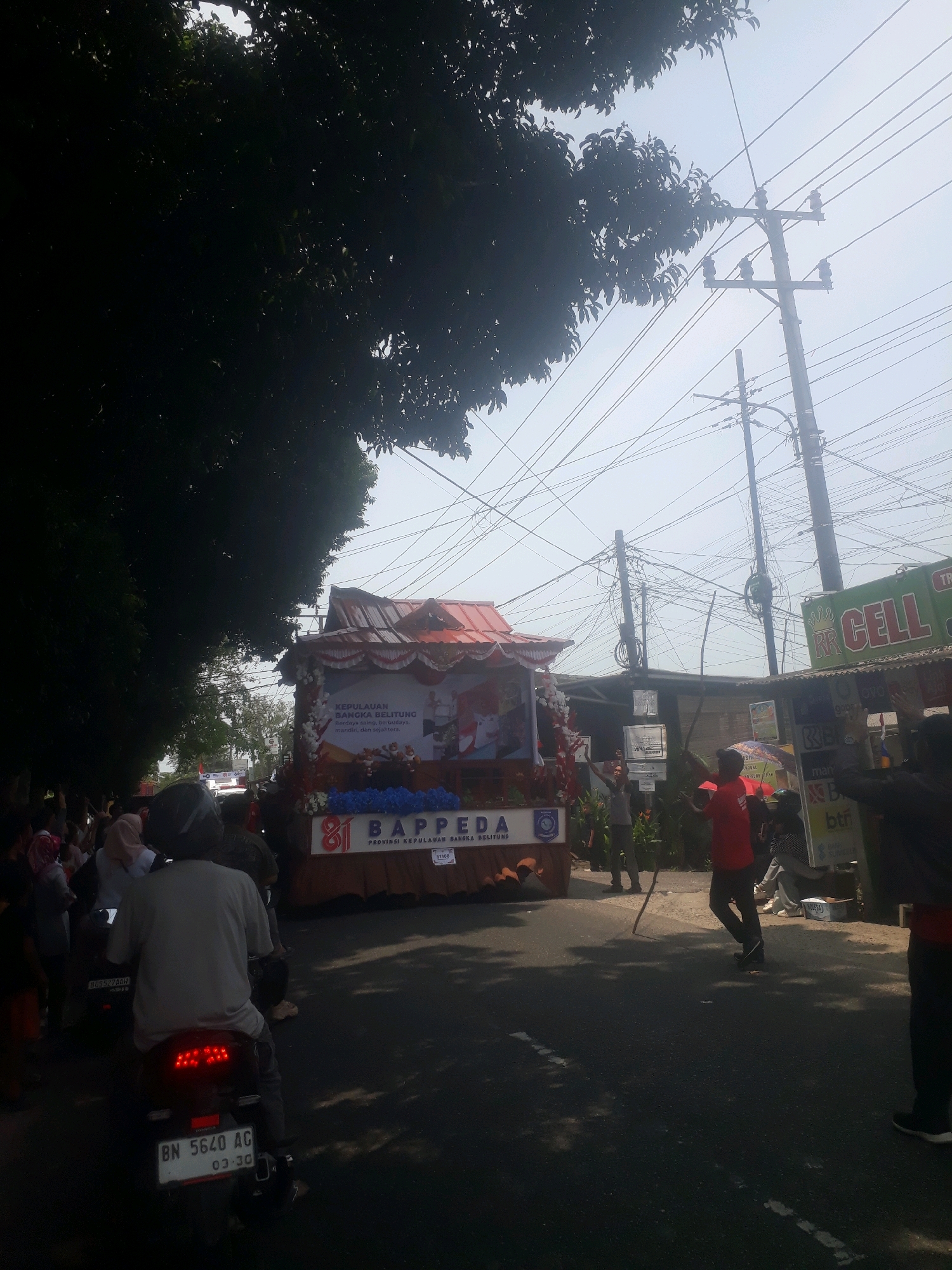 The image depicts a daytime street scene, likely a parade or festival, on Jalan Jenderal Sudirman in Pangkalpinang, Indonesia. A decorated float, possibly representing the Bangka Belitung Province (KEPULAUAN BANGKA BELITUNG) based on visible text, is the central focus. The float features red and white decorations and a large banner with text and imagery.
Several individuals are visible. In the foreground, a person wearing a grey shirt and a helmet is riding a motorcycle. Further back, on the right side of the street, a person in a red shirt is holding a stick and appears to be participating in the event. Other individuals are lining the street, suggesting an audience or participants in the parade.
The environment includes a paved road with white lane markings and shadows cast by trees and overhead structures. Electrical poles with numerous wires are prominent in the background, a common sight in urban Indonesian settings. A commercial establishment with a green sign reading "RR CELL" is visible on the right side of the street. The sky is overcast or hazy, indicating a bright but diffused light condition. The overall impression is of a lively public gathering in an urban environment.