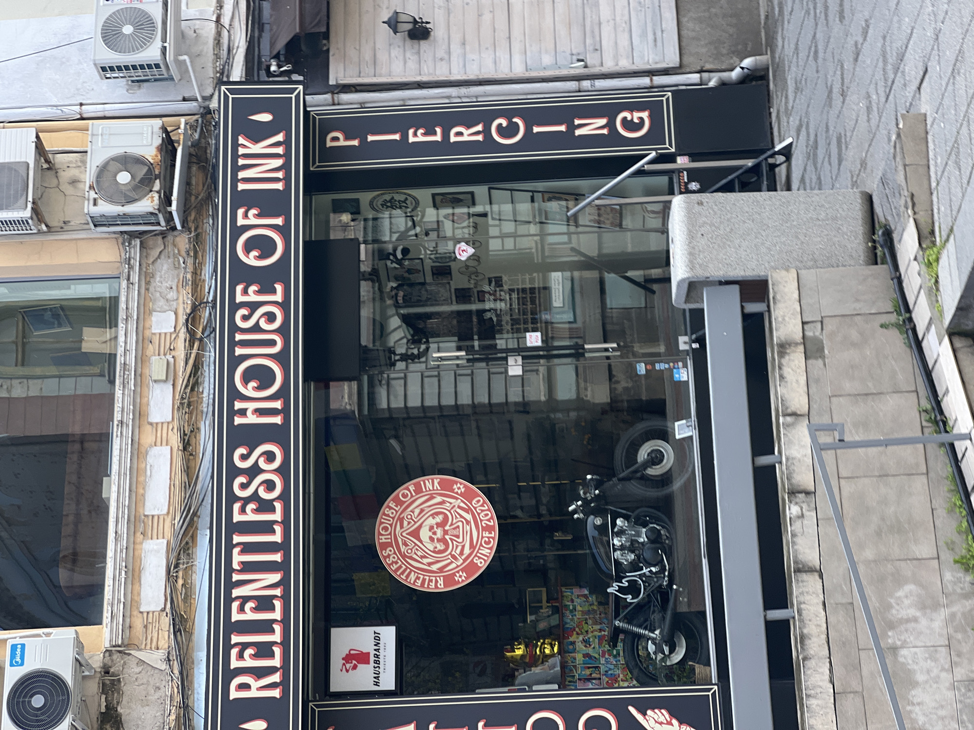 The image shows the facade of "Relentless House of Ink", a tattoo and piercing studio in Plovdiv, Bulgaria. The storefront features large signs with "RELENTLESS HOUSE OF INK" vertically aligned on one side and "PIERCING" horizontally above the entrance. A motorcycle is visible behind the glass. Air conditioning units are attached to the building's exterior. The sidewalk and a portion of the street are also visible.