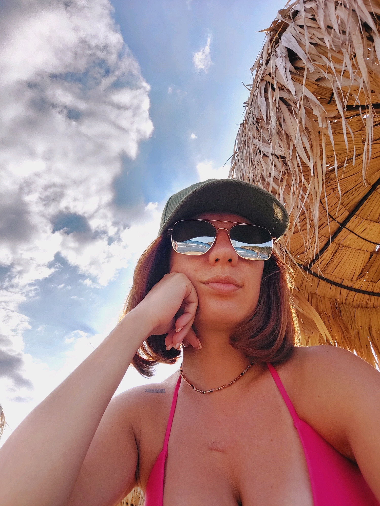 A woman is taking a selfie on a beach in Sozopol, Bulgaria. She is wearing a green cap, sunglasses, and a pink bikini. She is resting her chin on her hand and looking at the camera. The sky is blue with white clouds, and there is a straw umbrella above her. The reflection in her sunglasses shows a beach scene with people and buildings.