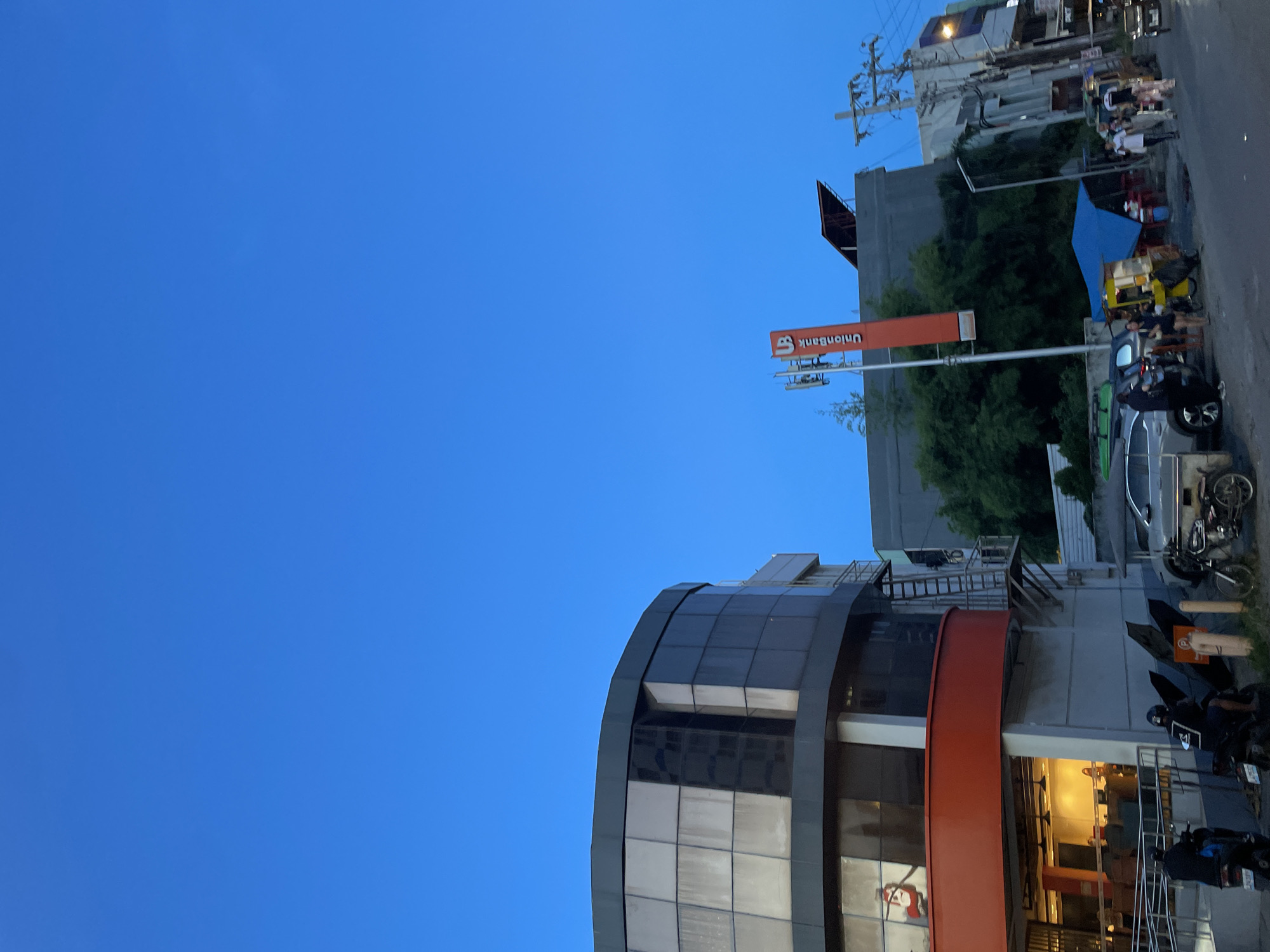 The image captures a street scene in Davao City, Philippines, under a clear blue twilight sky. A modern, multi-story building with a curved glass facade and an orange-colored lower level dominates the left side of the frame. Adjacent to it, a tall, orange sign with the "UnionBank" logo is visible. In the background, other buildings, including a gray structure with trees in front, and utility poles line the street. The street itself is paved and appears to be moderately busy, with several vehicles including cars, motorcycles, and a three-wheeled vehicle. A few people are seen walking and standing near the roadside, and some are on motorcycles. The overall atmosphere suggests an urban environment during the evening transition.