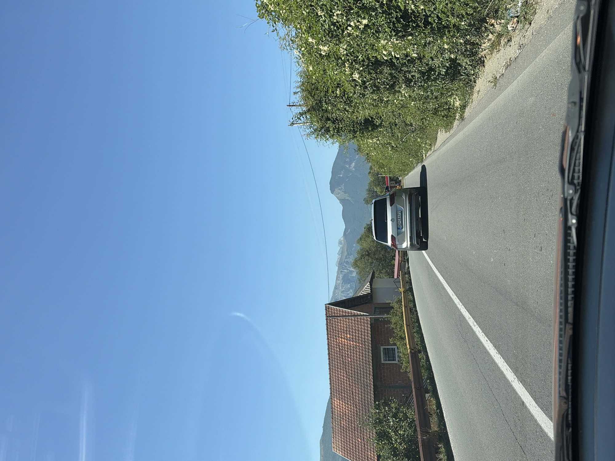 In the city of Martinaj, Montenegro, a silver Volkswagen Tiguan drives on an asphalt road under a clear blue sky. The car is heading away from the viewer, with its rear end visible. The road is marked with a white dashed line on the left and a solid white line on the right.

To the left of the road, a house with a reddish-brown tiled roof and brick walls is visible. The house has a small white-framed window and is surrounded by green foliage. A yellow metal barrier runs along the edge of the road in front of the house.

To the right of the road, a dense hedge of green bushes, some with white flowers, lines the path. In the distance, mountains with rugged peaks are silhouetted against the sky. The overall environment appears rural and natural, with a sense of open space and clear weather.

The image is captured from inside a vehicle, as indicated by the visible windshield wipers and the top edge of the dashboard at the bottom of the frame. The scene is well-lit, suggesting it was taken during daylight hours.