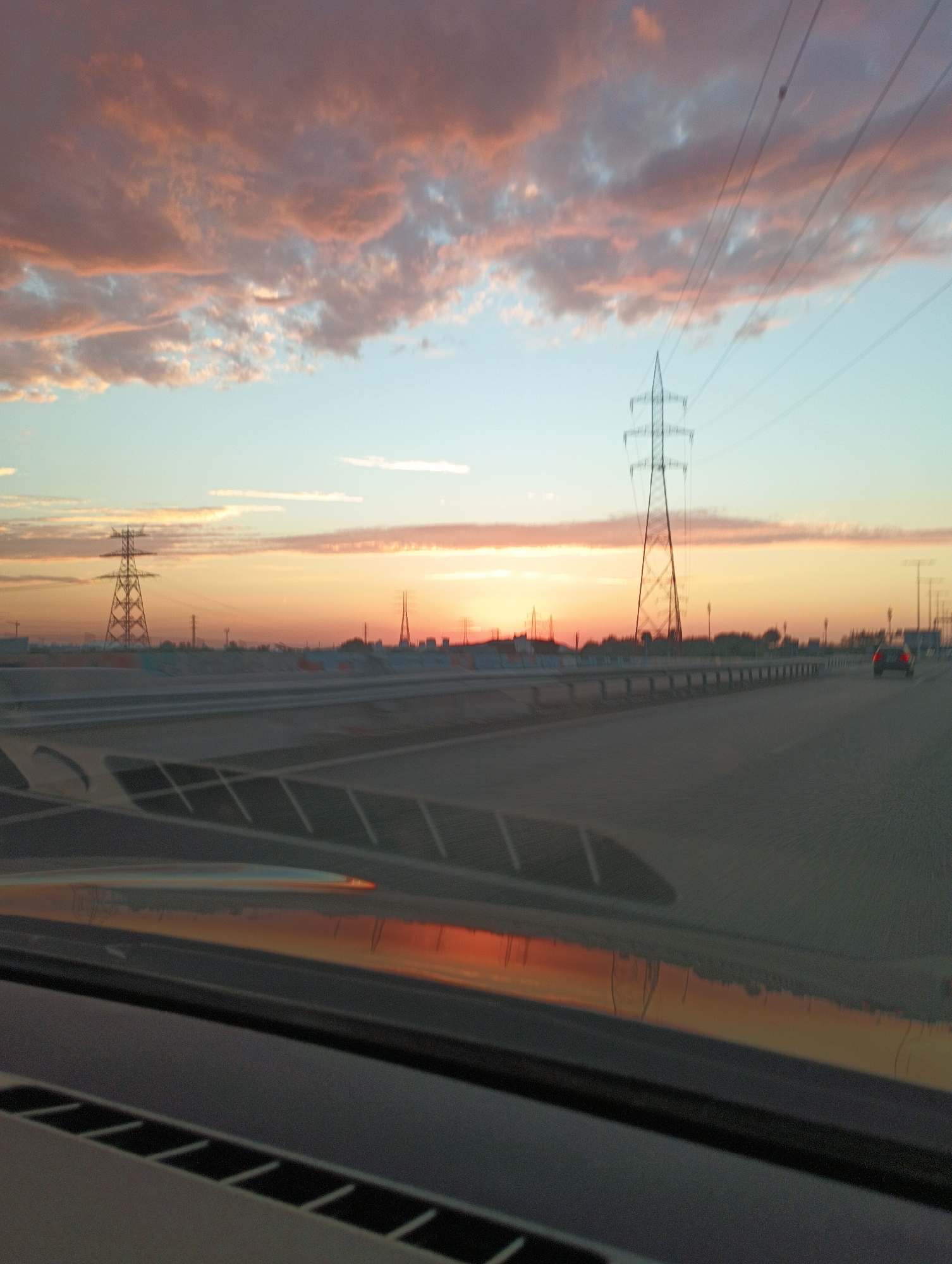 This is a medium shot, eye-level photograph taken from inside a vehicle looking out at a sunset scene. The sky is a gradient of colors, from a pale blue at the top to a fiery orange and pink near the horizon. Clouds are scattered across the sky, tinged with pink and orange from the setting sun. In the distance, there are several power line towers, one of which is particularly prominent in the center of the frame. The towers are silhouetted against the bright sky. A highway or road stretches across the middle ground, with a car visible driving away from the viewer on the right side of the road. The foreground of the image shows the interior of the vehicle, with the dashboard and windshield visible, reflecting the sunset colors. The overall impression is of a scenic drive at dusk.