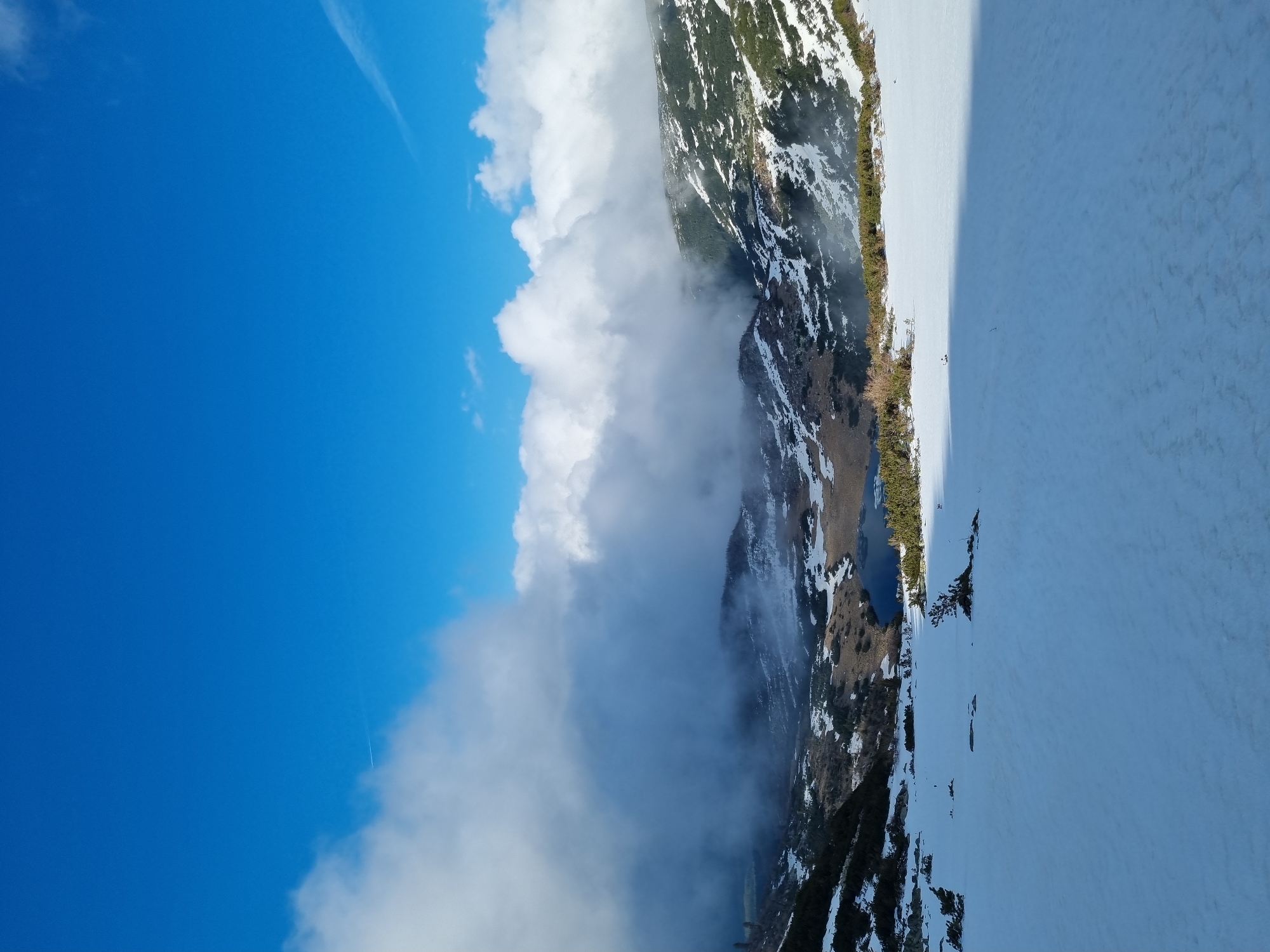 The image depicts a landscape in Govedartsi, Bulgaria, featuring a mountain range partially obscured by low-lying clouds on its left. A clear blue sky is visible above the clouds. On the right, a snow-covered area extends, possibly a slope or valley floor. A small, dark blue lake is nestled within the landscape, surrounded by patches of vegetation and snow. The composition is vertically oriented.