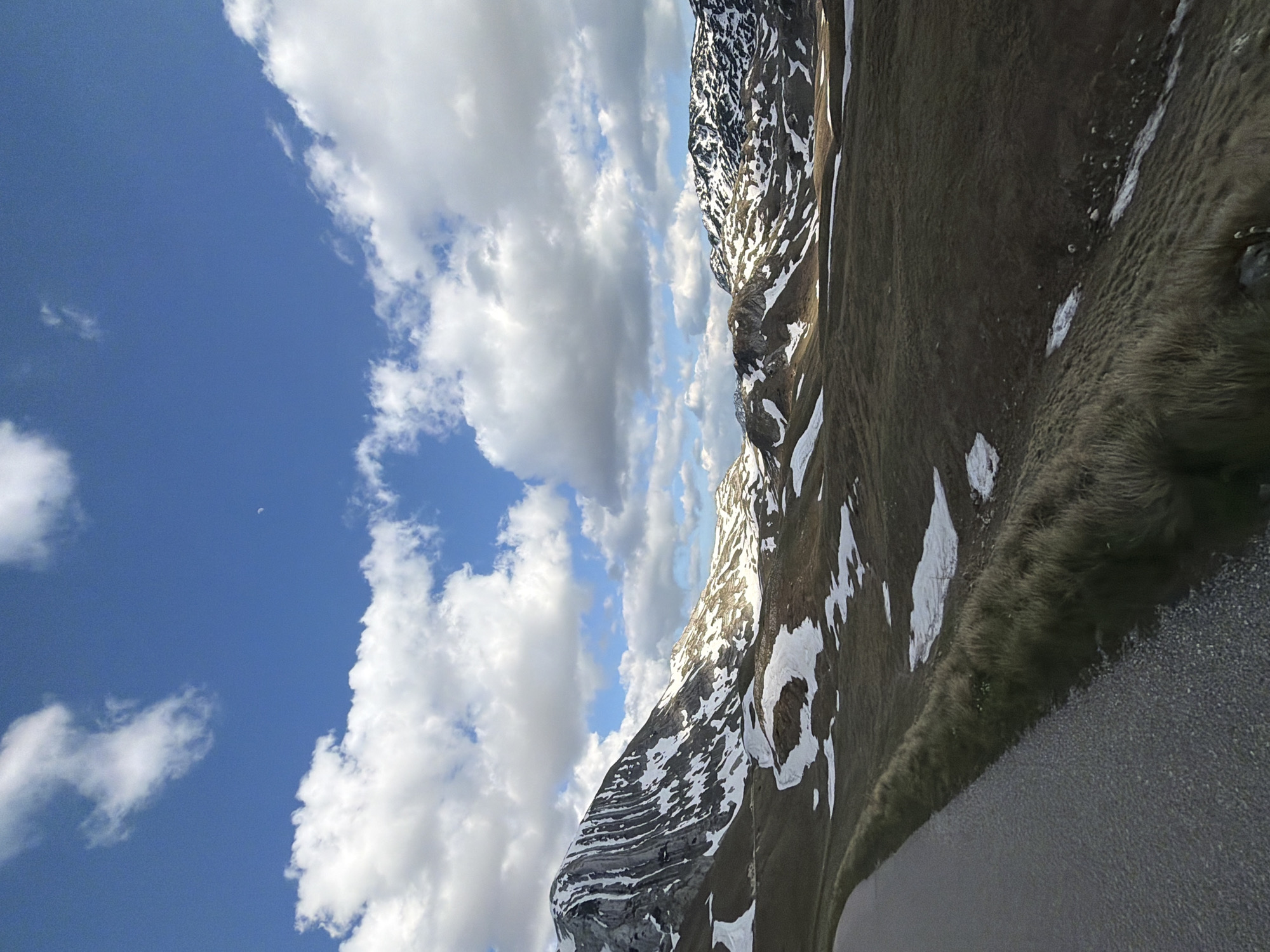 The image depicts a landscape in Боричје, Montenegro. There are mountains with patches of snow on them. The terrain is covered in grass and a paved road is in the foreground. The sky is blue with white, puffy clouds. A small, faint crescent moon is visible in the blue sky.