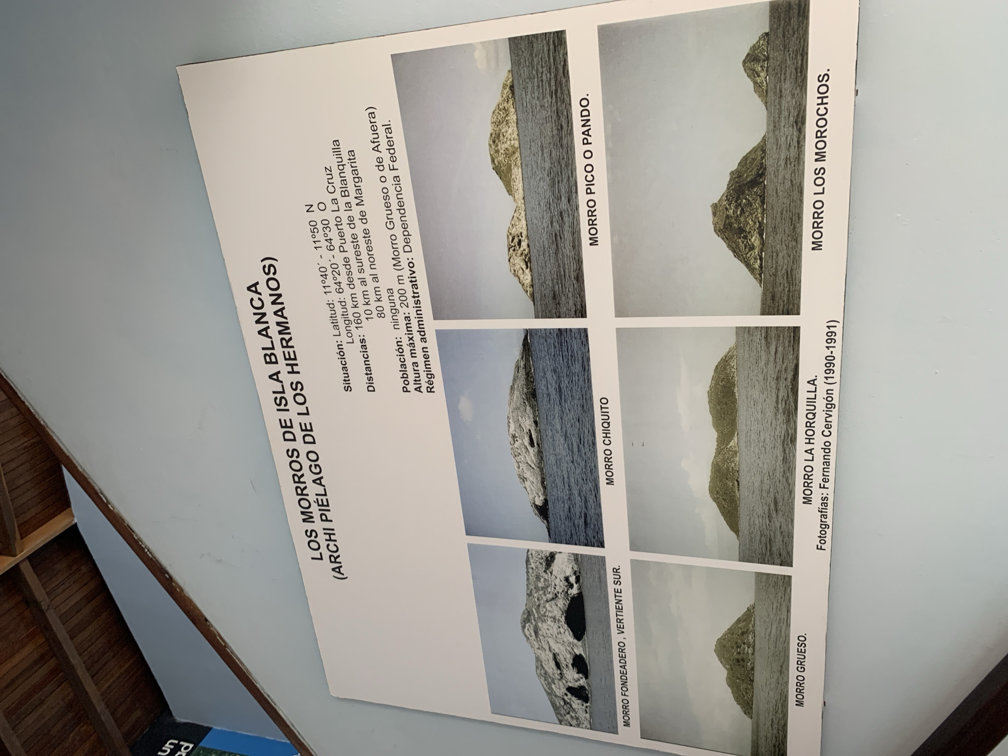 The image is a photograph of a sign or informational display about "LOS MORROS DE ISLA BLANCA (ARCHI PIÉLAGO DE LOS HERMANOS)". The sign is mounted on a light blue wall and has a wooden ceiling structure above it. The display features a title, geographical information (latitude, longitude, distances, population, maximum altitude, administrative region), and six photographs of different rock formations or small islands in the sea. The text describes the location as being in Boca del Río, Venezuela. The photographs are labeled with names such as "MORRO FONDEADERO, VERTIENTE SUR", "MORRO CHIQUITO", "MORRO PICO O PANDO", "MORRO GRUESO", "MORRO LA HORQUILLA", and "MORRO LOS MOROCHOS". The bottom of the sign also states "Fotografías: Fernando Cervigón (1990-1991)". The overall presentation suggests an educational or tourist-oriented display.