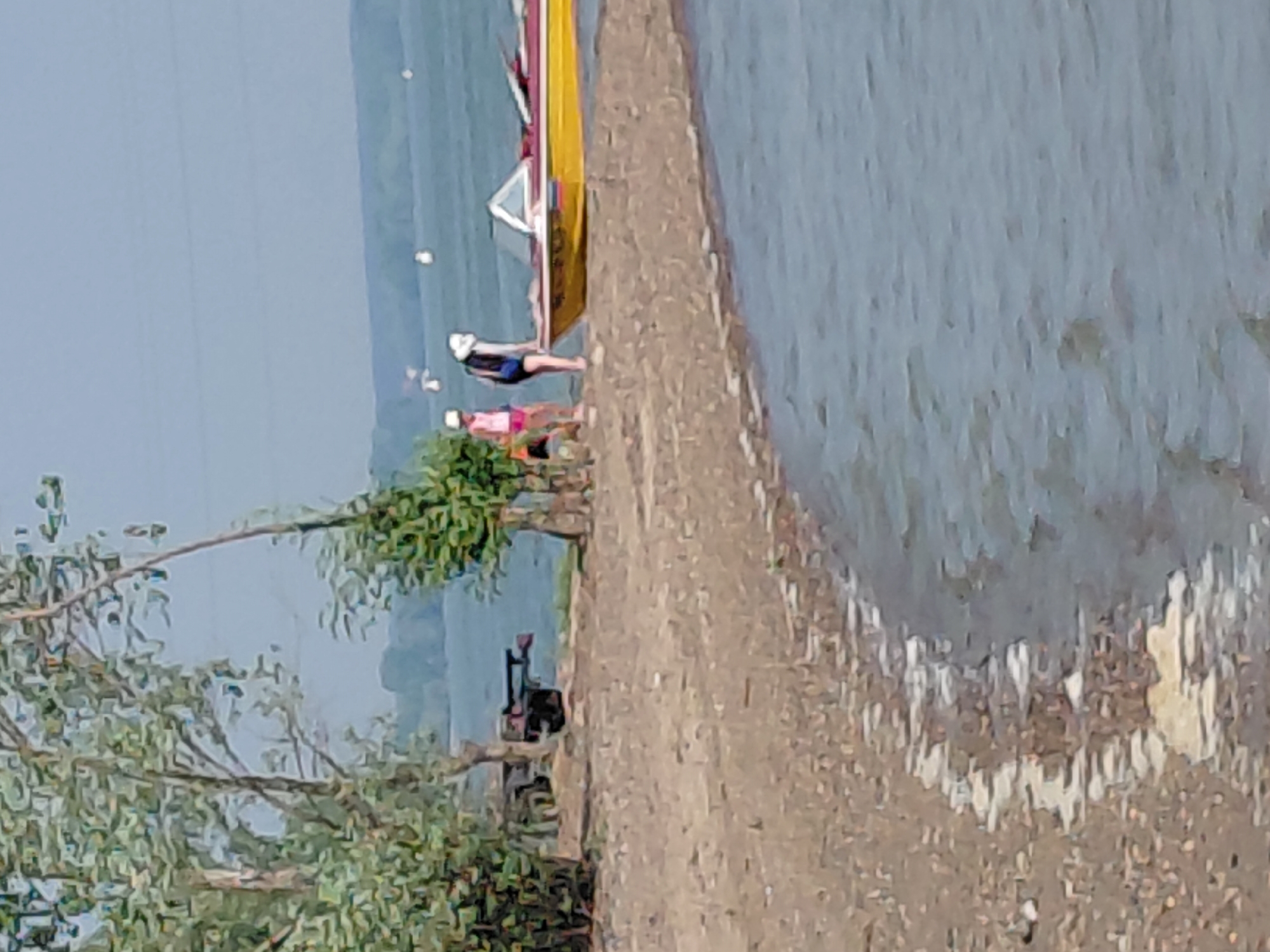 This media file depicts a scene along the Ob River in Novosibirsk, Russia. The image is oriented vertically, showing a waterfront area. In the foreground, there is a concrete embankment and some foliage.
Several people are visible. One person, wearing a white cap, dark shorts, and a dark top, is lying on their back on the embankment, appearing to be sunbathing or relaxing. Another person, in a pink top and darker shorts, is partially visible nearby. Further along the embankment, a dark object, possibly a piece of equipment or furniture, is present.
A prominent yellow and red object, likely a boat or a part of a boat, is leaning against the embankment. The water of the Ob River fills the right side of the frame, with ripples suggesting a calm or slightly breezy condition. The background shows a hazy landscape with a pale blue sky.
The overall impression is of a casual, recreational activity taking place outdoors, possibly during the daytime with fair weather. The scene suggests a moment of leisure by the river. No text is visible in the image.