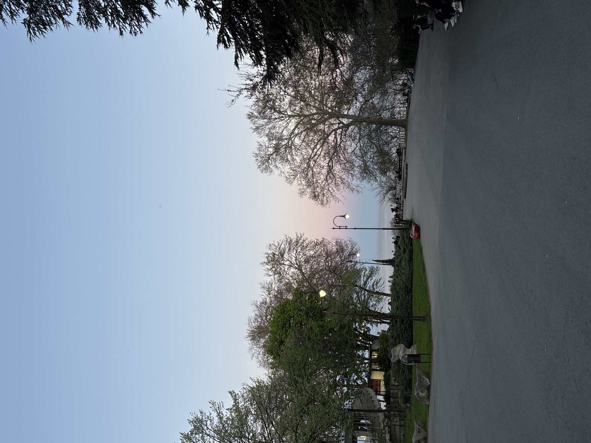 The image depicts a view of the Varna Sea Garden in Varna, Bulgaria. A paved pathway runs along the right side of the frame, lined with trees, bushes, and streetlights. On the left, the sky transitions from blue to pink, indicating either sunrise or sunset. Several trees, some with bare branches and others with green leaves, frame the sky. Silhouettes of people can be seen along the coastline in the distance. At the top right, a partial view of people's shoes and legs suggests a crowd.