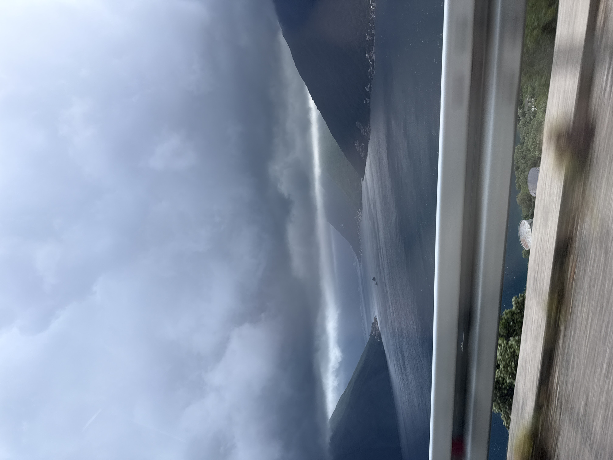 The image shows a fjord landscape under a dark, cloudy sky, likely taken from a moving vehicle. A bright ray of sunlight pierces through the clouds, illuminating the water and a section of the steep, mountainous terrain on either side of the fjord. A guardrail is visible in the foreground, suggesting the viewpoint is from a road or highway. The blurred scenery outside the window implies the vehicle is in motion. Considering the city and country provided, this scene is highly likely to be in or around the Bay of Kotor, specifically in Montenegro. The photograph does not show any people, subjects or interactions. The environment is a cloudy day over a fjord, as viewed from inside a moving vehicle.
