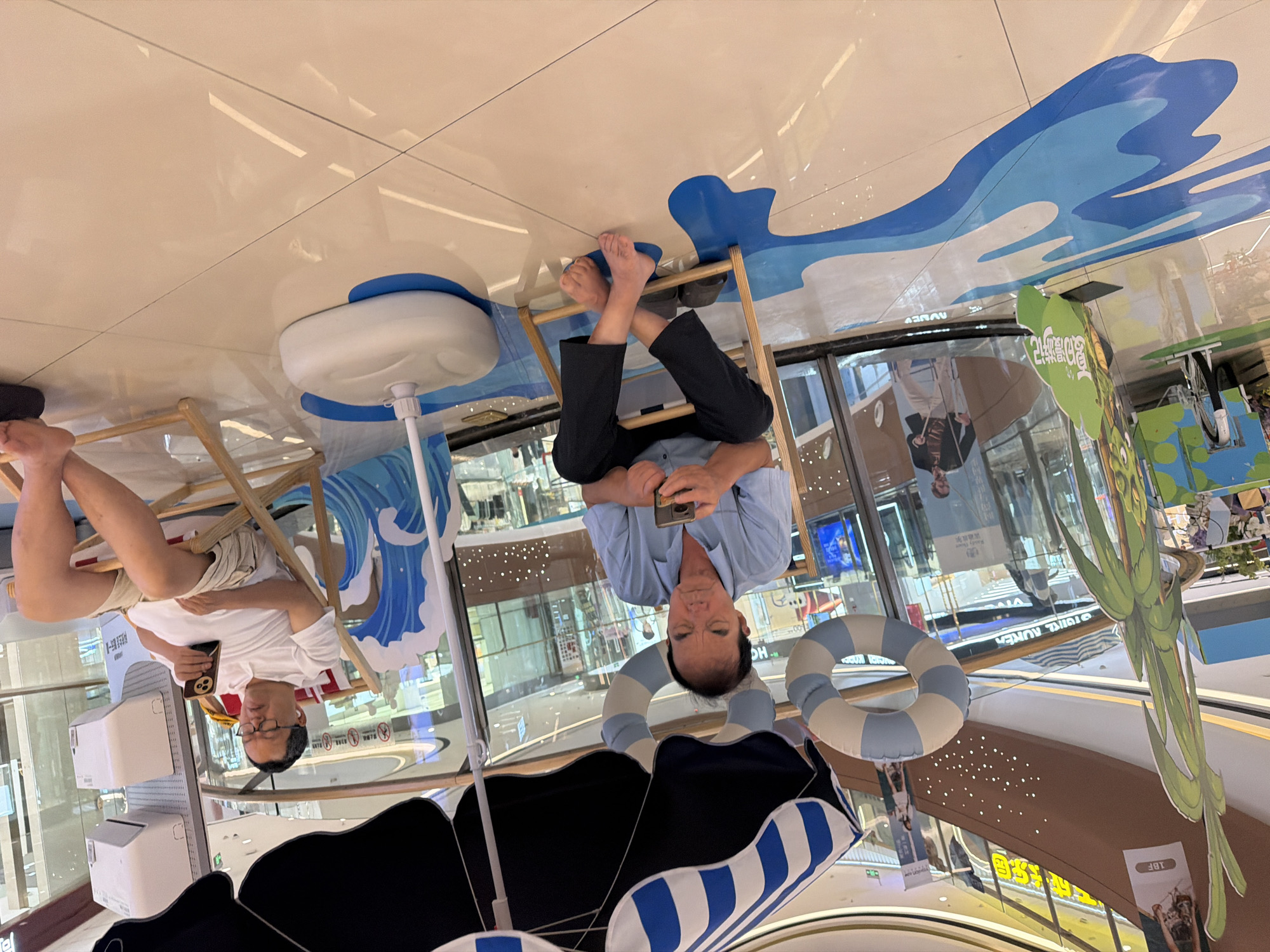 This image captures a scene within a shopping mall in Jiangjin, China, featuring two men relaxing under a large umbrella that is decorated with blue and white stripes. The floor is tiled with a reflective, beige surface, and features a blue and white wavy design painted on it, simulating water.
In the foreground, an older man with dark hair is seated in a wooden deck chair. He is wearing a light blue collared shirt and dark trousers. He is looking down at his mobile phone, holding it in his hands, with his legs crossed. His feet are bare. Next to his chair is a dark-colored pair of slippers.
To the right, another man is seated in a similar wooden deck chair. He is wearing glasses, a white polo shirt, and light-colored shorts. He is also looking at his mobile phone, which he holds in his right hand. He has white earbuds in his ears.
Behind the men, there are decorative elements suggesting a beach or summer theme, including life preservers hanging from the umbrella structure and a cutout of blue waves. In the background, the mall environment is visible, with shops and signage. To the left, a large green cutout resembling a palm tree stands near the edge of the scene. There is also a bicycle parked behind the palm tree cutout.