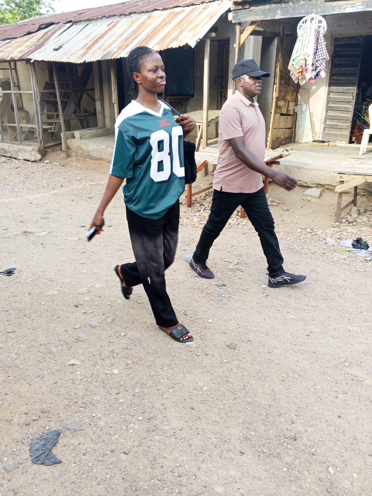 The image depicts two individuals walking on a dirt road in what appears to be a residential area of Adankolo, Nigeria. The road is sparsely covered with pebbles and debris.
A woman wearing a green and white sports jersey with the number 80 on it, along with black pants and sandals, is walking towards the left of the frame. She is holding a mobile phone in her left hand.
To her right, a man wearing a light brown polo shirt, black pants, a black cap, and sneakers is walking in the same direction.
The background includes buildings with corrugated metal roofs, some with open fronts or unfinished construction. Clothes hang on a line attached to one of the buildings. Simple wooden furniture can also be seen nearby.