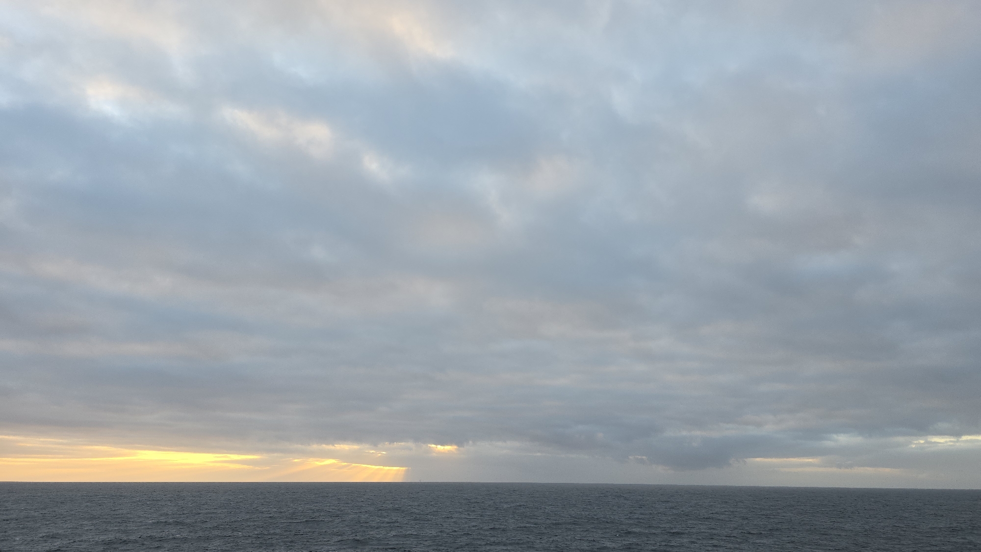 The image shows a seascape under a cloudy sky. The sky is predominantly gray with patches of light, possibly indicating the location of the sun near the horizon. The sea is dark and calm with minimal visible waves. The horizon line is straight, with the transition from sea to sky marked by a band of light reflecting off the water. There are no people, objects, or subjects visible other than the sea and the sky. The location in the city is Unknown, Unknown.