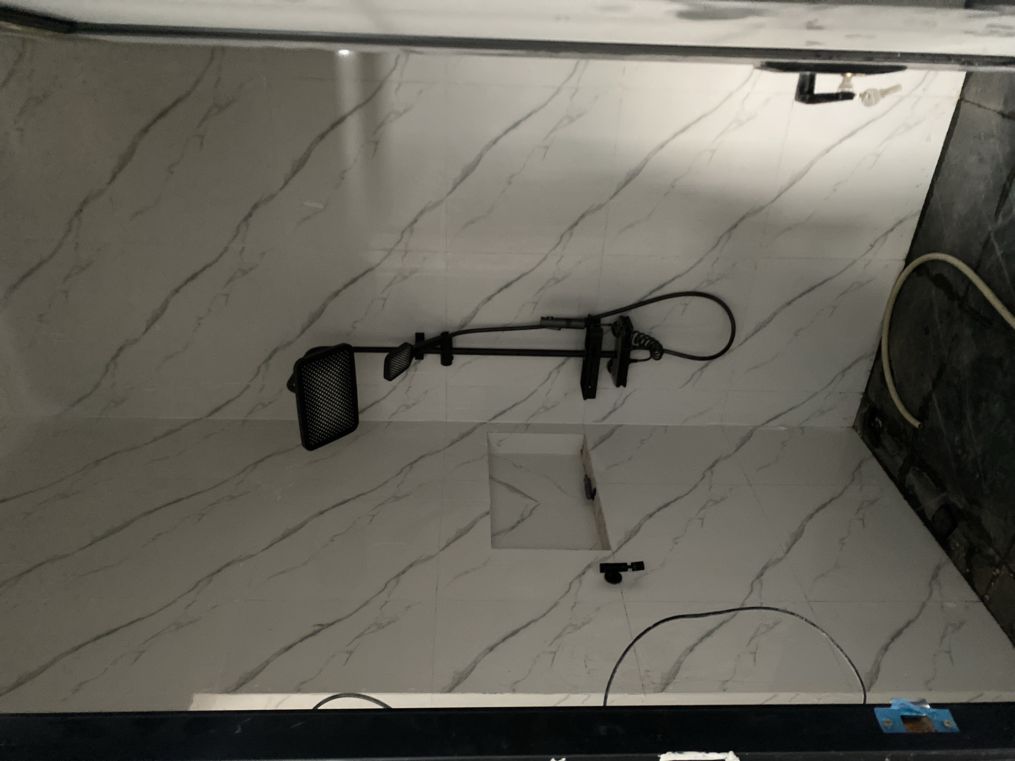 This media file depicts a bathroom, likely in a residential setting in Cottrueng, Indonesia. The walls are tiled with a white marble-patterned finish, featuring prominent grey veining. A black showerhead and hose assembly is mounted on the wall. Below the shower, there is a rectangular recessed area in the tiled wall, possibly for storing toiletries or as a niche. A small, dark fixture is visible on the tiled floor, to the right of the recessed area. A portion of a light-colored hose and a dark, possibly black, coiled cord are also present on the floor. The overall impression is of a modern bathroom under construction or recently renovated, given the exposed elements and the presence of a coiled cord. The lighting suggests it's daytime or an interior setting with artificial lighting. There are no people visible in the image.