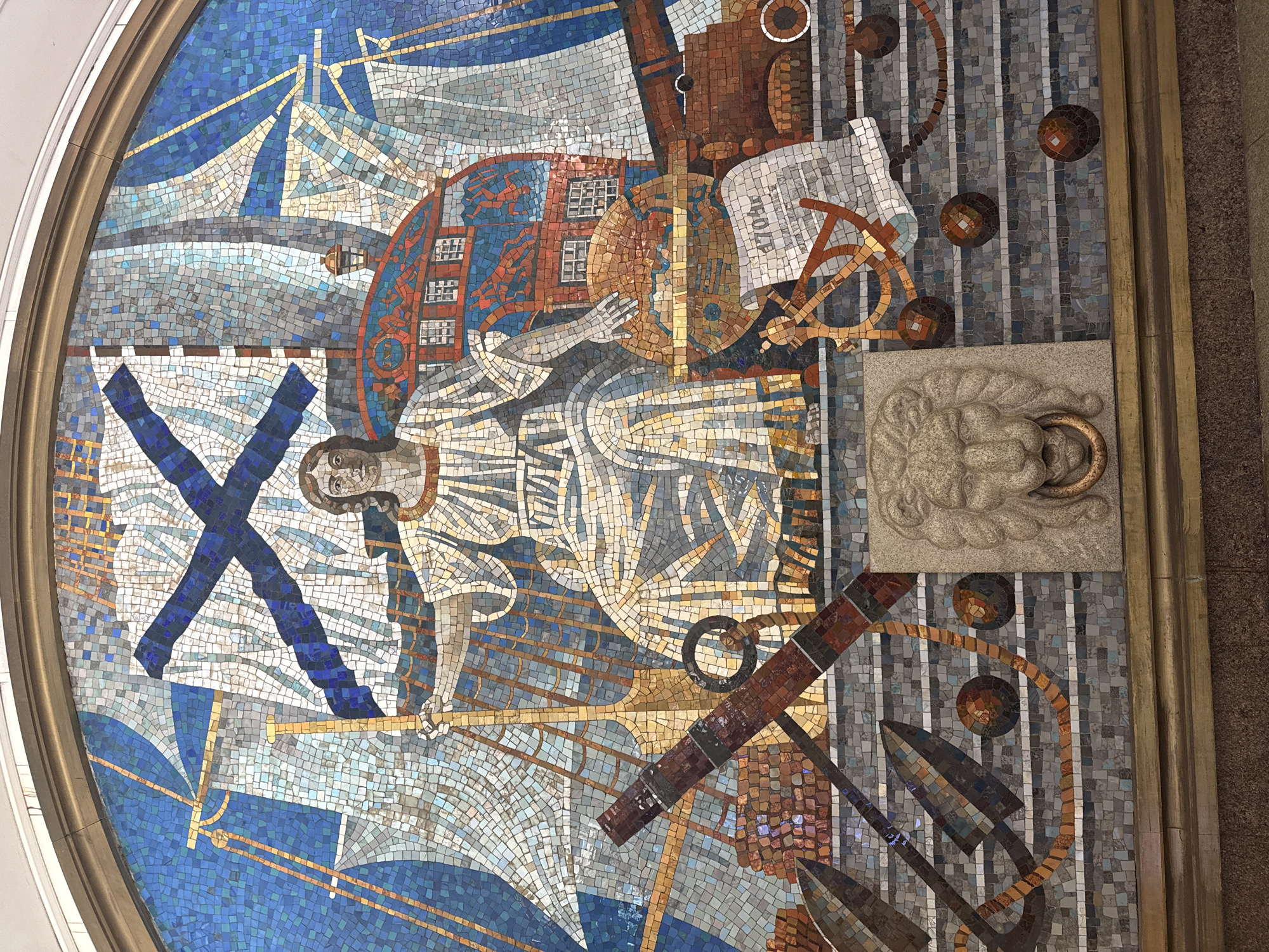 This is a mosaic mural in the lobby of the Admiralteyskaya metro station in Saint Petersburg, Russia. The mural depicts various nautical and historical symbols.
A central figure appears to be a woman or an allegorical figure representing navigation or the sea. She is surrounded by elements such as a ship with sails, represented by blue, gray, and white tiles. A blue cross (possibly the St. Andrew's Cross, a symbol of the Russian Navy) is prominently featured on the left side.
Other elements include an anchor, a lion's head sculpture with a ring in its mouth, a building, a telescope, circles, and other nautical instruments and decorative elements. The mosaic is made of small, colorful tiles, creating a detailed and intricate design.
