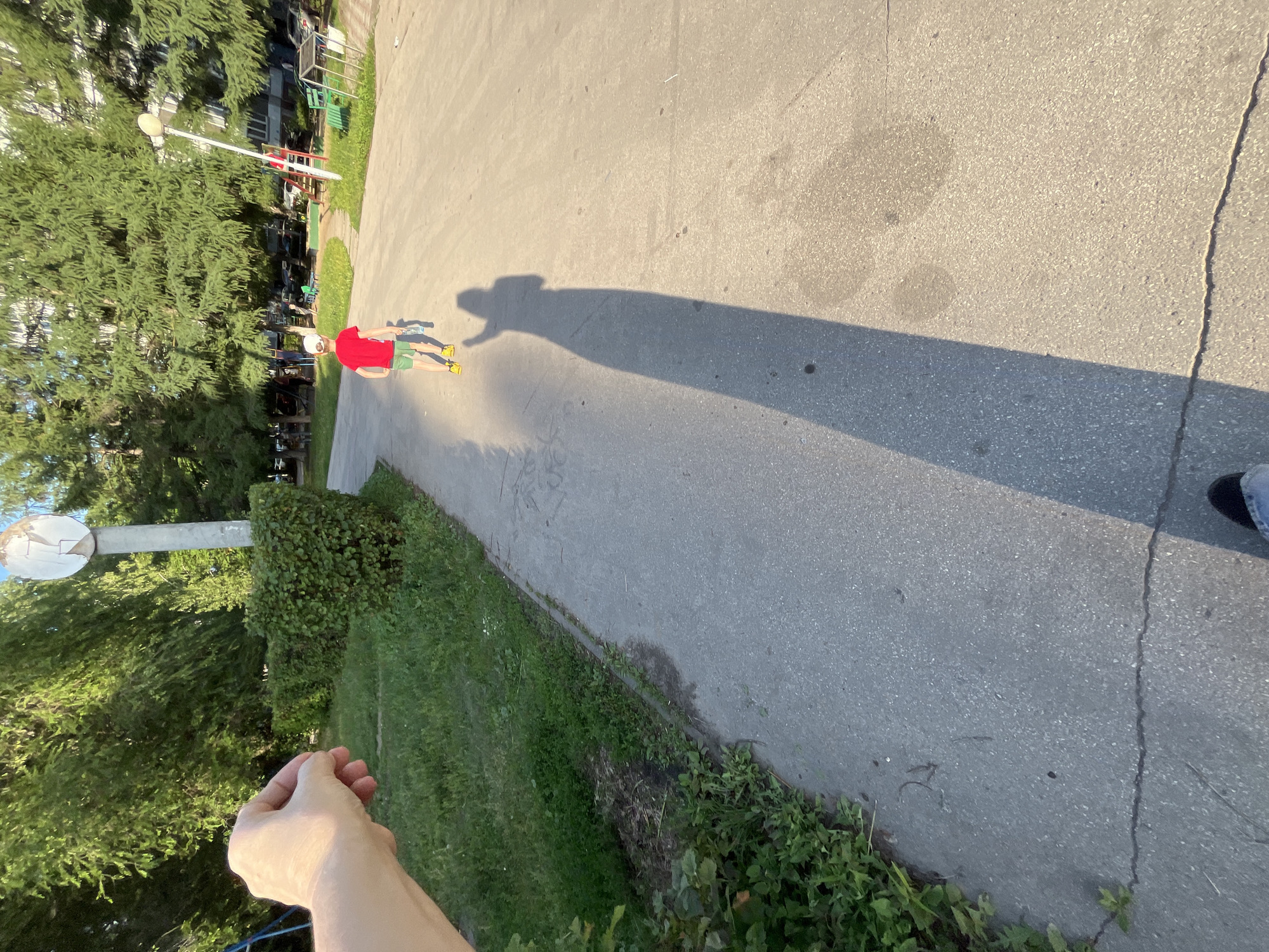In a sunlit, outdoor urban environment, a young boy wearing a red t-shirt, green shorts, and a white baseball cap walks away from the camera. He is holding a bottle in his right hand. The boy is on a paved walkway that curves to the right. A long shadow of a person extends across the walkway and onto the grass to the left. In the foreground, a person's left hand and forearm are visible, reaching out towards the scene. To the left of the walkway is a patch of green grass with some bushes and a tall pole with a damaged circular sign. In the background, tall trees form a canopy over the area, and beyond the trees, apartment buildings are visible. A playground with a slide and swings can be seen to the right of the walkway. The overall scene appears to be a residential courtyard or park.