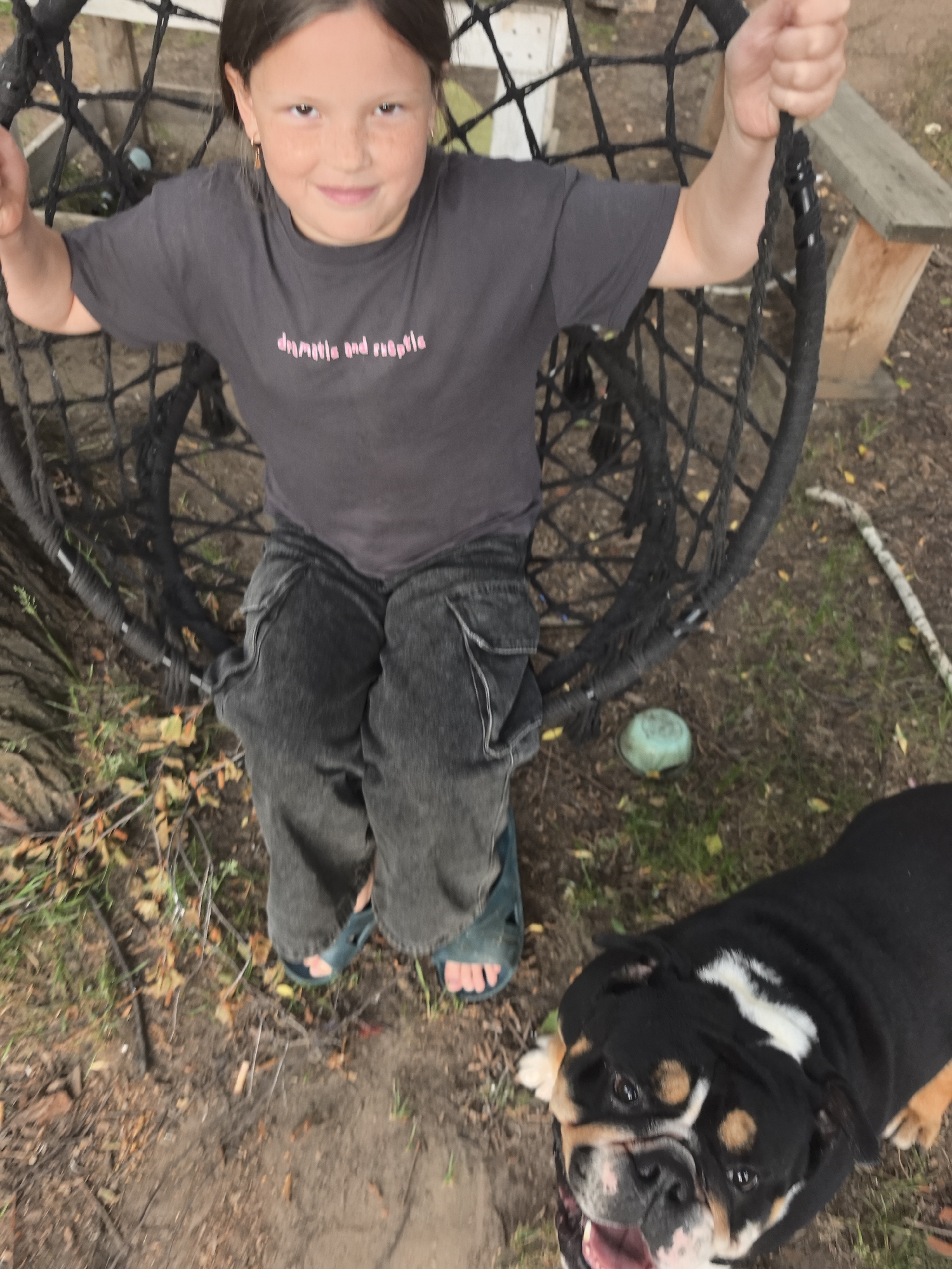 A young girl with brown hair and freckles is sitting in a black net swing. She is wearing a dark grey t-shirt with pink text and dark grey cargo pants. She is smiling and looking at the camera. Next to her, on the ground, is a black and white bulldog with its mouth open. The background appears to be an outdoor setting with dirt, fallen leaves, and some wooden structures. The specific location in Noviy, Russia, is not discernible from the image.