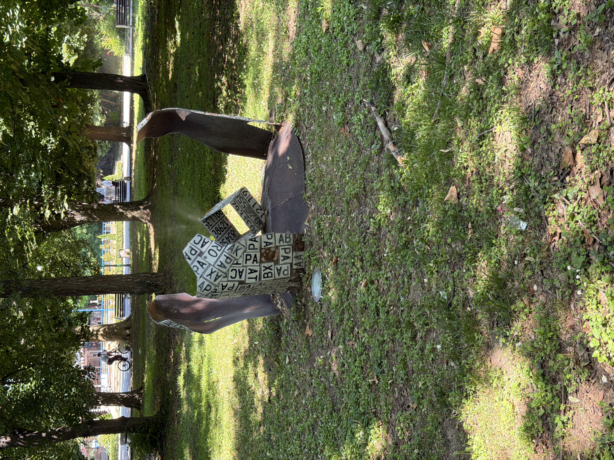 This image depicts an outdoor scene in the city of Pazardzhik, Bulgaria. In the foreground, a metallic sculpture is situated on a grassy area with scattered fallen leaves and small plants. The sculpture is abstract, consisting of curved metallic elements and geometric shapes adorned with text and numbers, possibly resembling license plates. A small circular light fixture is embedded in the ground near the sculpture's base.
In the middle ground, a paved path runs through the park, bordered by trees with dense green foliage. A person is seen riding a bicycle on this path, and another individual is walking near a park bench. In the background, more trees and park infrastructure, including benches and lampposts, are visible. The overall environment suggests a public park setting during daytime, with sunlight filtering through the tree canopy, creating shadows on the ground.
