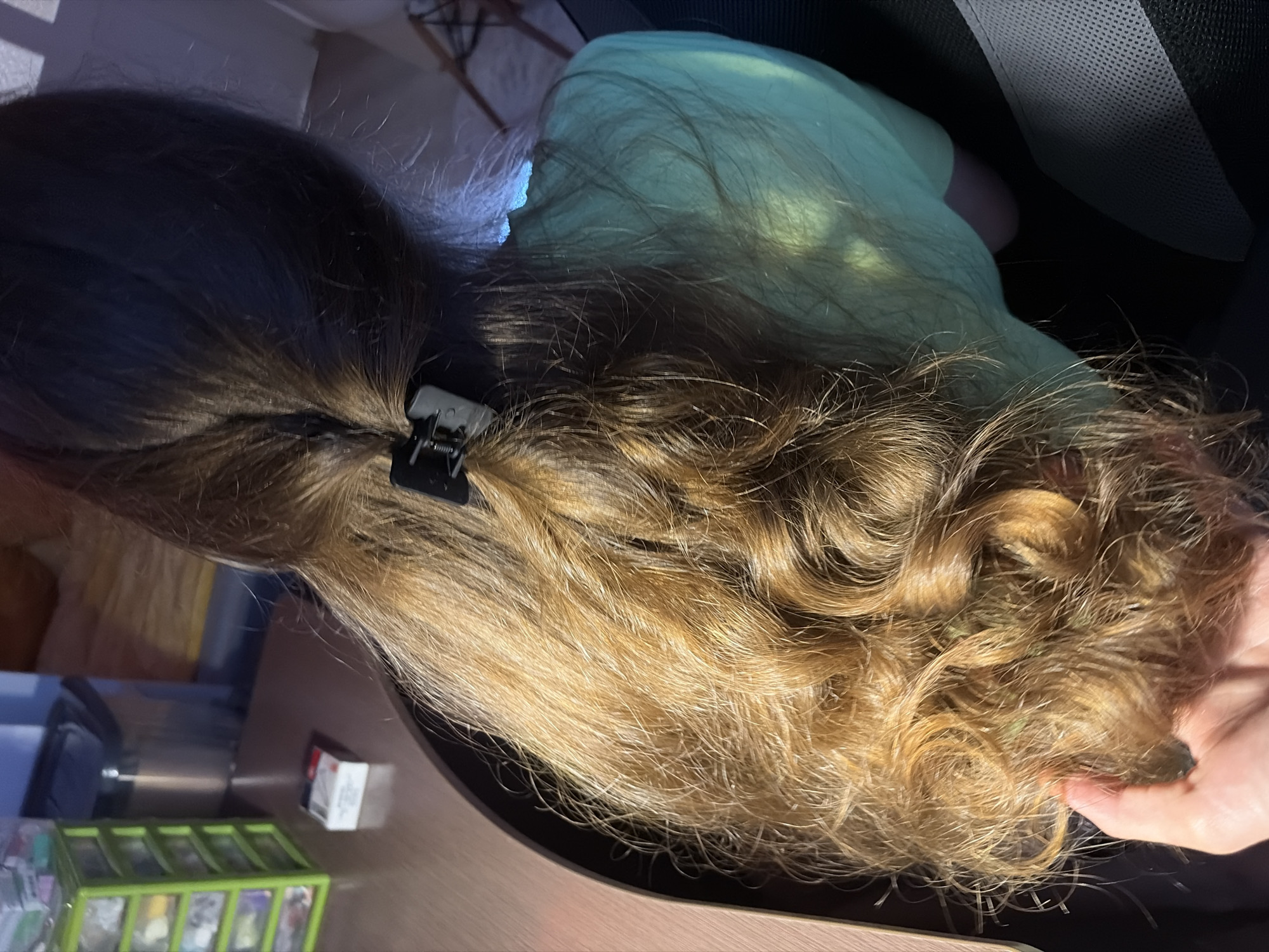 The image shows a close-up, overhead view of a person's long, wavy, light brown hair. The hair is partially pulled back with a black hair clip. The person is wearing a light green t-shirt. A hand is visible in the lower left corner, gently touching the hair. The background includes a wooden desk with a small stack of white and red packaging, and a green plastic organizer with drawers. The lighting suggests an indoor environment, possibly at night, with artificial light sources casting shadows. The scene does not provide any specific indicators of its location within Leninsk-Kuznetskiy, Russia.