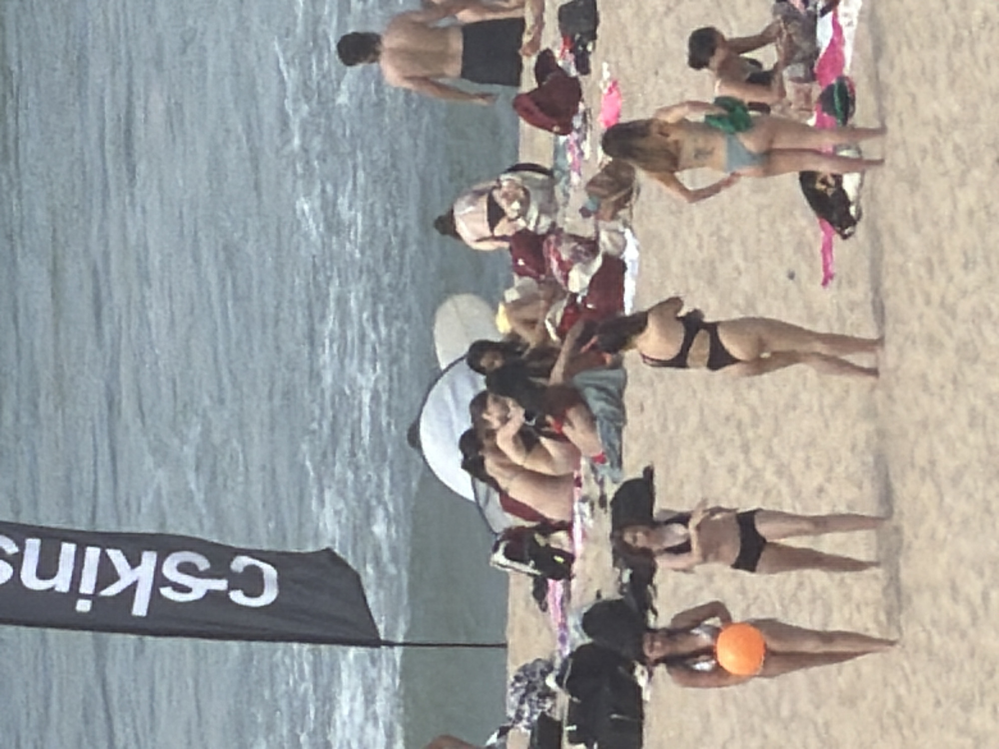 A medium-shot, full-body photograph depicts a beach scene in Espinho, Portugal, with several individuals enjoying the day by the ocean. In the foreground, two women are walking on the sand. The woman on the left, facing left, is wearing a silver bikini and holding an orange ball. The woman on the right, facing forward, is wearing a black bikini with shorts.
Further back, a group of people are seated on the sand near the water. One woman in a light-colored bikini is facing away from the camera, bending over. Several other individuals are visible in the background, some lying down and others seated. A dark flag with white lettering, possibly a brand logo, stands on a pole to the left of the frame. The ocean with gentle waves forms the backdrop to the sandy beach.