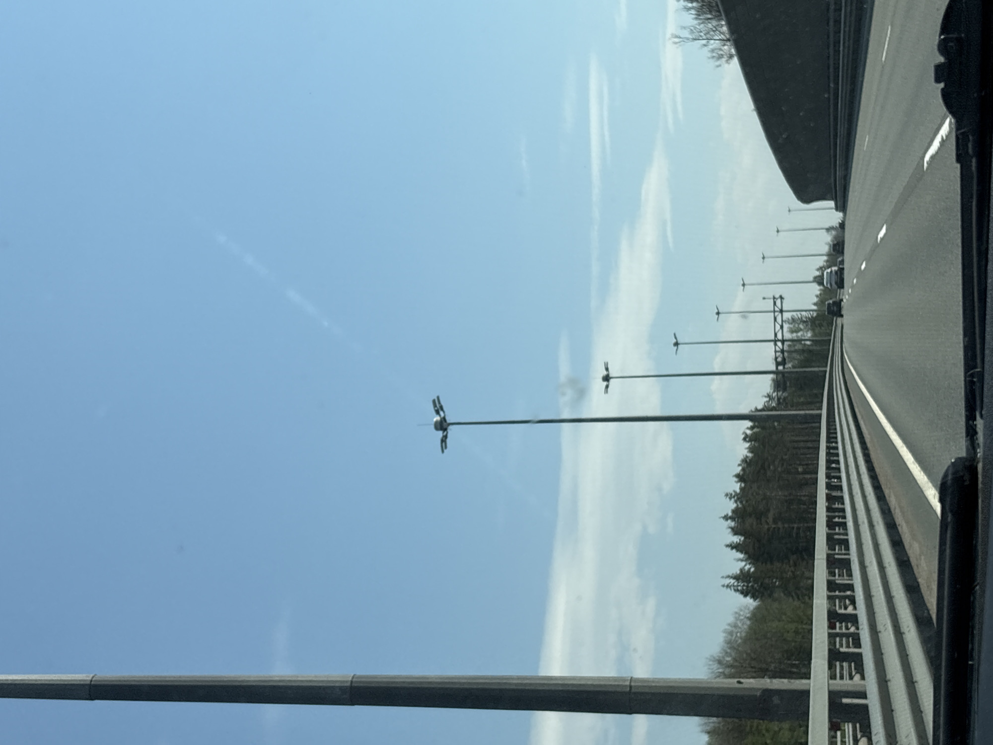 The image shows a highway scene in Pesochny, Russia, viewed from the perspective of a vehicle moving along the road. The sky is blue with wispy clouds. Rows of highway light poles line the road. A barrier runs along the edge of the highway, separating the road from a forested area. Cars can be seen in the distance.
