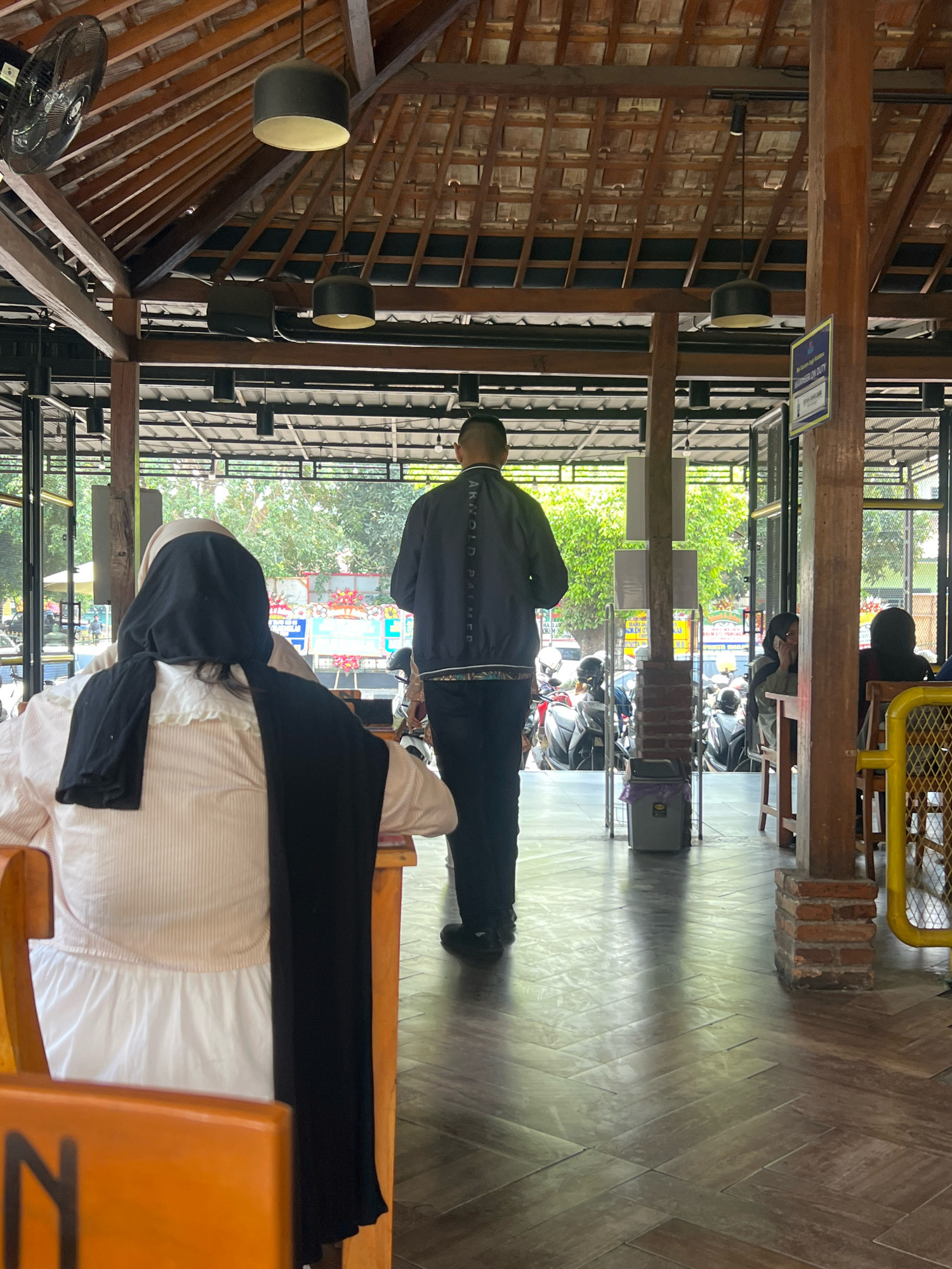 The image depicts the interior of an open-air establishment with a wooden roof structure, likely a restaurant or cafe, in Yogyakarta, Indonesia. A person from behind, wearing a light-colored striped shirt and a black headscarf, is seated at a wooden table in the foreground. To their left, a part of an orange chair with a black letter "N" is visible. In the midground, a person wearing a dark jacket with lettering on the back is standing and facing away from the camera, looking towards an outdoor area. Beyond them, through glass panels, is a street scene with parked motorcycles and trees. To the right, another individual is seated at a table and speaking on a cell phone, with a yellow metal structure and more wooden furniture visible. The floor is tiled in a herringbone pattern, reflecting the natural light streaming in. Brick pillars support the wooden roof. A trash can with a purple bag is visible near the entrance to the outdoor area. The overall atmosphere is casual and outdoorsy.