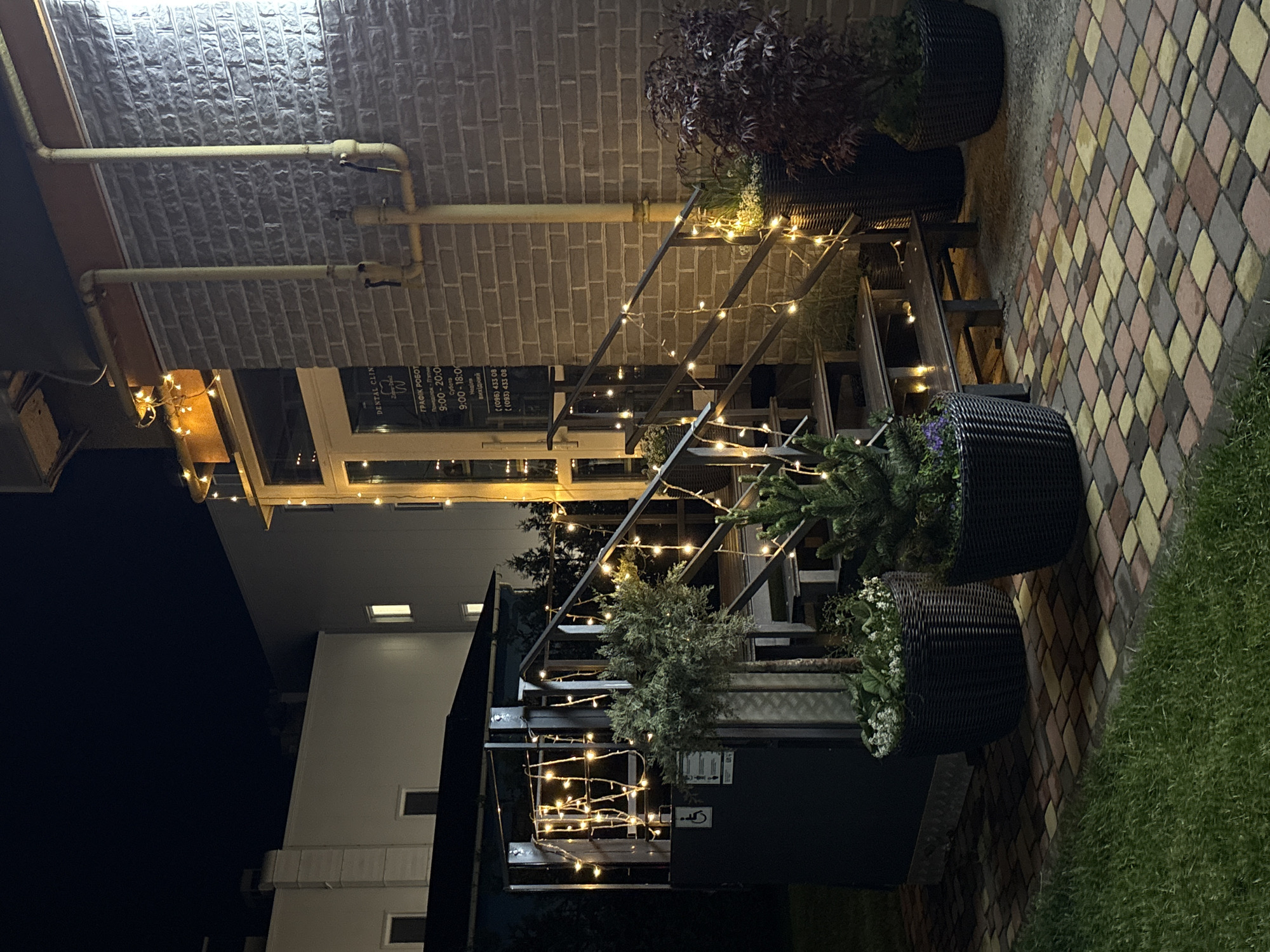 An outdoor night-time view of a building in Hatne, Ukraine, captures a welcoming entrance area. The building's facade on the left features an upper section of light grey brickwork with yellow utility pipes running across it, and a lower section of smooth, light-colored walls with large windows or glass doors.
A metal outdoor staircase, adorned with numerous warm, glowing string lights, leads up to the building's entrance. Along the staircase and on adjacent surfaces, several dark wicker planters hold a variety of plants, including evergreens, reddish foliage, and some with small white flowers, adding to the decorative appeal.
The ground area to the right of the building consists of colorful patterned paving stones in shades of yellow, brown, and pink, transitioning to a patch of vibrant green grass further to the right. No people are visible, but the scene is brightly illuminated, suggesting an operational business or a thoughtfully lit exterior.
On one of the glass doors at the entrance, a sign for a "DENTAL CLIN" (Dental Clinic) is clearly visible. Below it, a "Work Schedule" (ГРАФІК РОБОТ) is displayed in Ukrainian, indicating operating hours: Monday-Friday from 9:00-20:00, Saturday from 9:00-18:00, and Sunday as a "day off" (вихідний). Two Ukrainian phone numbers, beginning with (096) and (093), are also listed. The overall impression is one of a clean, inviting, and well-maintained professional establishment.