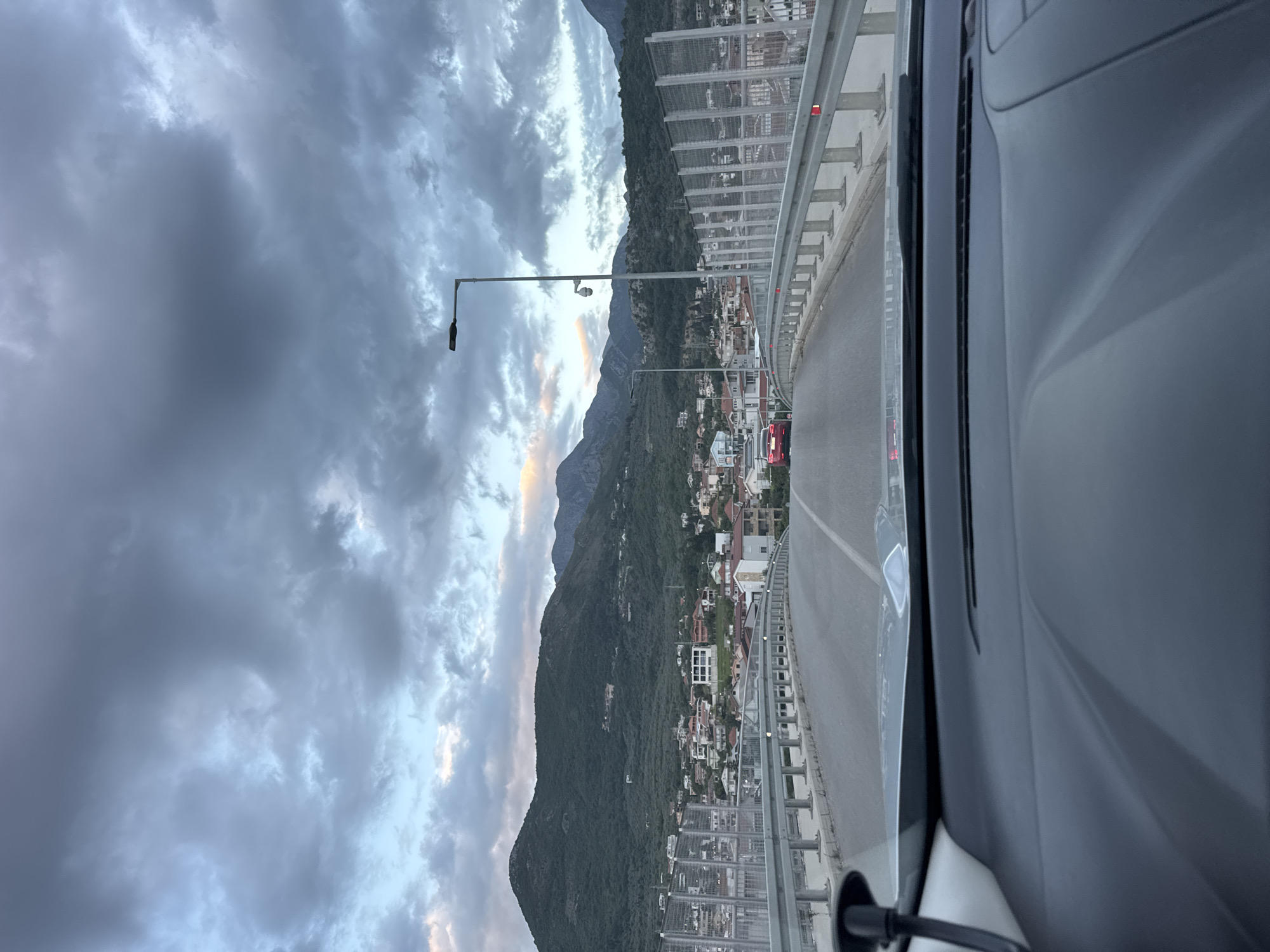The image, taken from inside a car, shows a view from the road on the bridge in Bar, Montenegro. The road bends to the right. Guardrails are visible on both sides of the bridge. Below the bridge, a densely populated area with residential buildings stretches towards a mountainside covered in lush green vegetation. The sky above is filled with heavy, dark clouds. A streetlight stands to the left of the road.