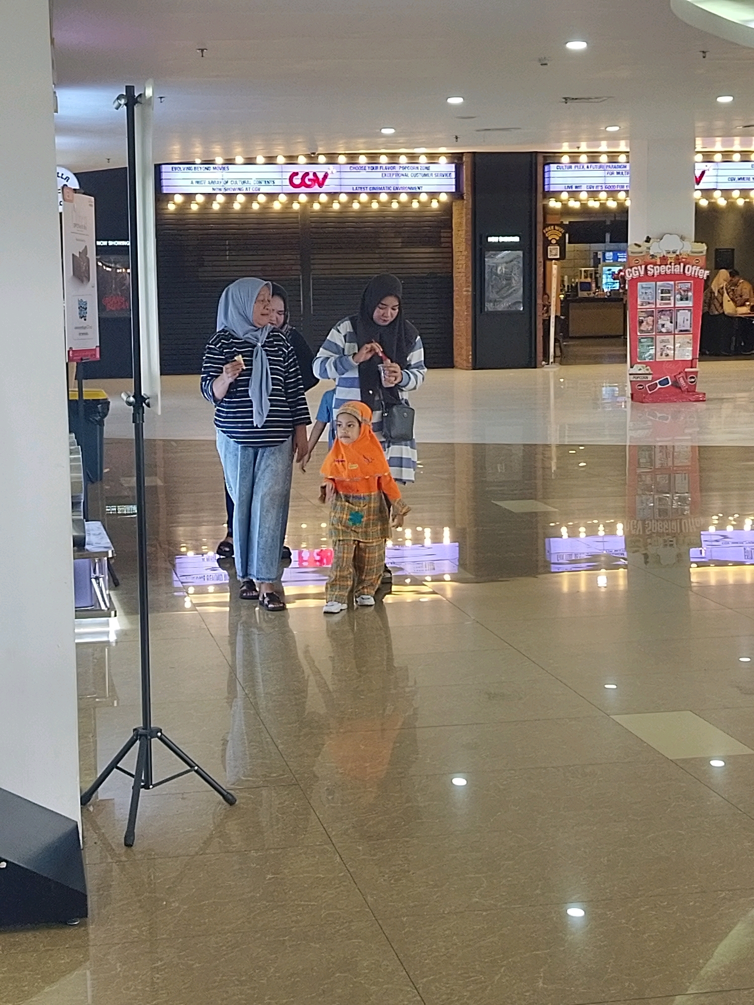 The image shows an indoor scene, likely a shopping mall or cinema lobby, in Mojokerto, Indonesia. In the foreground, a child wearing an orange hooded jacket and plaid pants, along with two adults, are walking. One adult, wearing a light blue headscarf and a striped shirt, is looking towards the left and holding a snack. Another adult, wearing a dark headscarf and a striped sweater, is walking behind her. The background features a movie theater entrance with illuminated signs and posters. A display for "CGV Special Offer" is visible on the right. The floor is polished and reflects the lights.